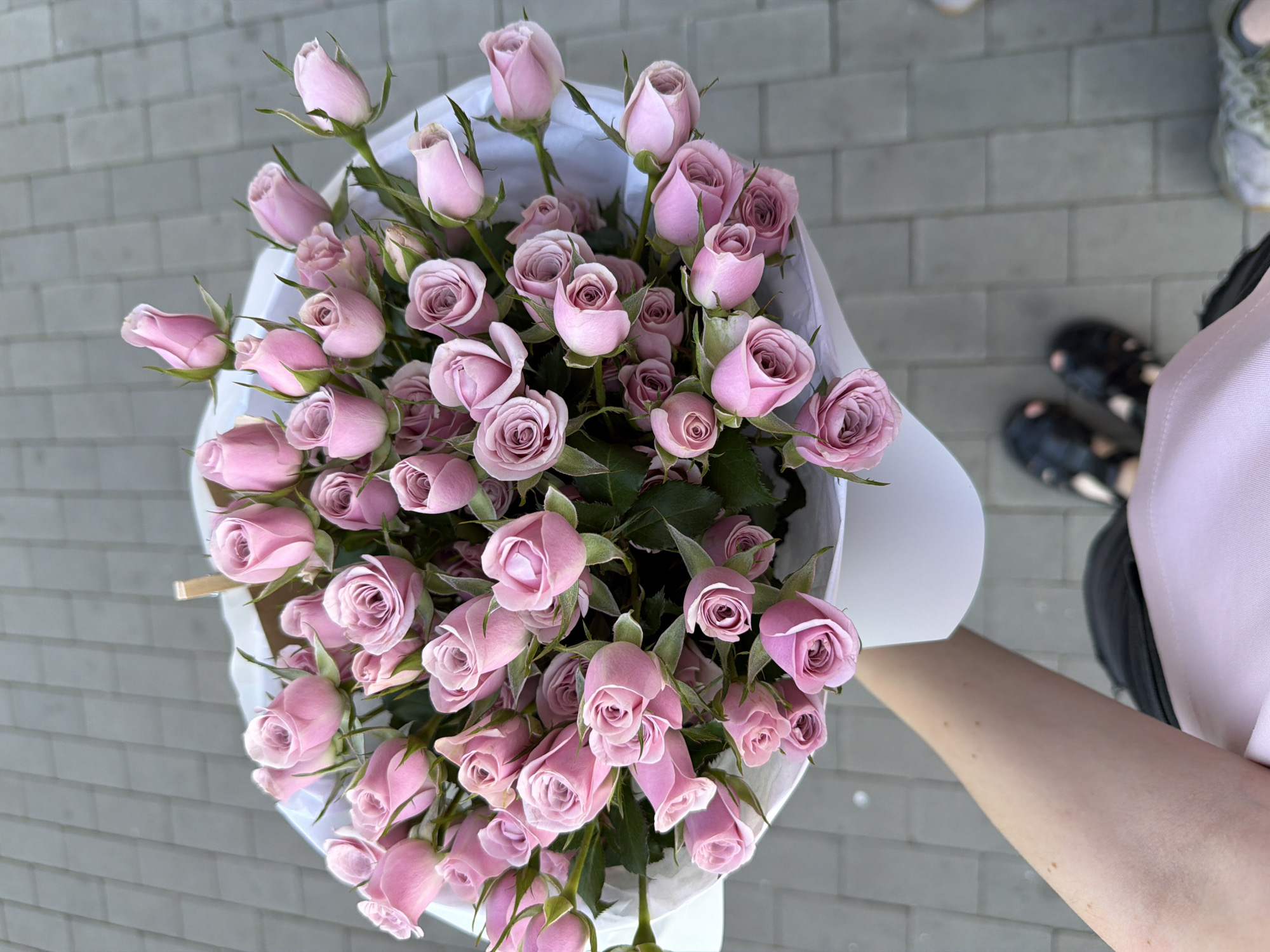 An overhead medium shot shows a hand holding a large bouquet of pink roses wrapped in white paper, positioned in front of a grey brick pathway. The roses are mostly closed buds with some slightly open, revealing lighter pink interiors. The bouquet is held in the lower left quadrant of the frame, obscuring some of the person's arm and shoulder. The hand is lightly tanned with a few brown spots. The person is wearing a pink garment. In the background, the pathway is made of rectangular grey bricks. Two pairs of feet are visible at the bottom of the frame; the left pair wears black open-toed shoes with pink embellishments, and the right pair is blurred and appears to be wearing patterned shoes. The scene is outdoors, possibly in a public space within the city of Kyiv, Ukraine.