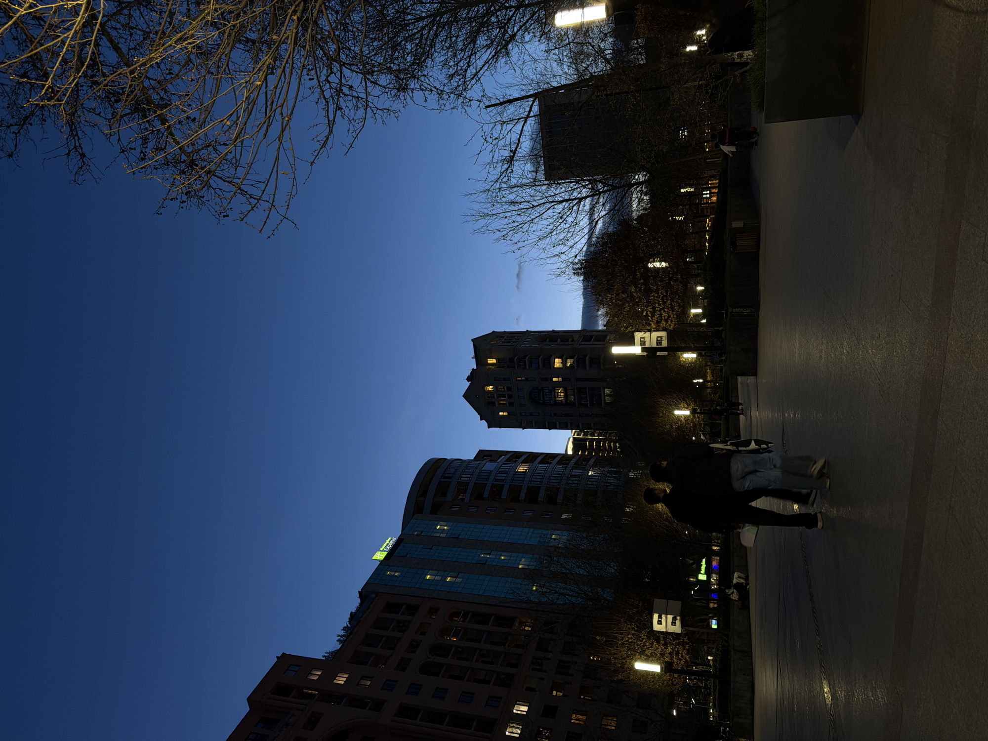 The image shows a nighttime scene in Yerevan, Armenia, featuring a paved area with several tall buildings visible against a dark blue sky. Tree branches, mostly bare, are visible at the top of the frame. Streetlights illuminate the walkway. Two figures appear to be walking in the middle of the scene. The Holiday Inn Express Yerevan hotel building is visible in the lower half of the image.