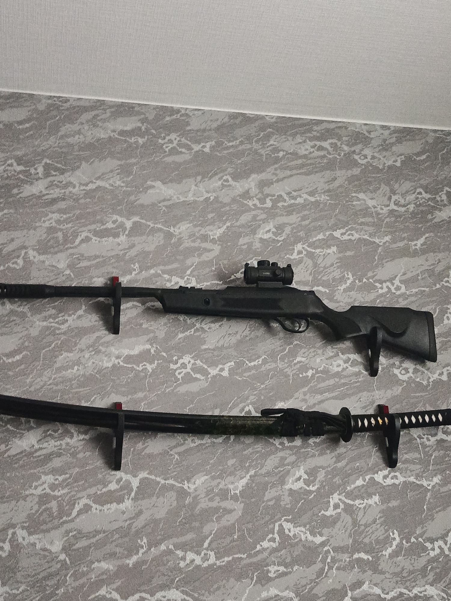 The image shows two weapons mounted on a wall with a marble-patterned wallpaper. The upper weapon is a black rifle with a scope. The rifle is mounted horizontally on a black bracket with a red accent. The lower weapon is a katana, also mounted horizontally on a black bracket with a red accent. The katana has a black handle and a decorative scabbard. The wall appears to be in an indoor setting. There are no people visible in the image.
The provided context states that the image is from the city of Lesosibirsk, Russia, but this information cannot be independently verified from the visual content of the image itself.