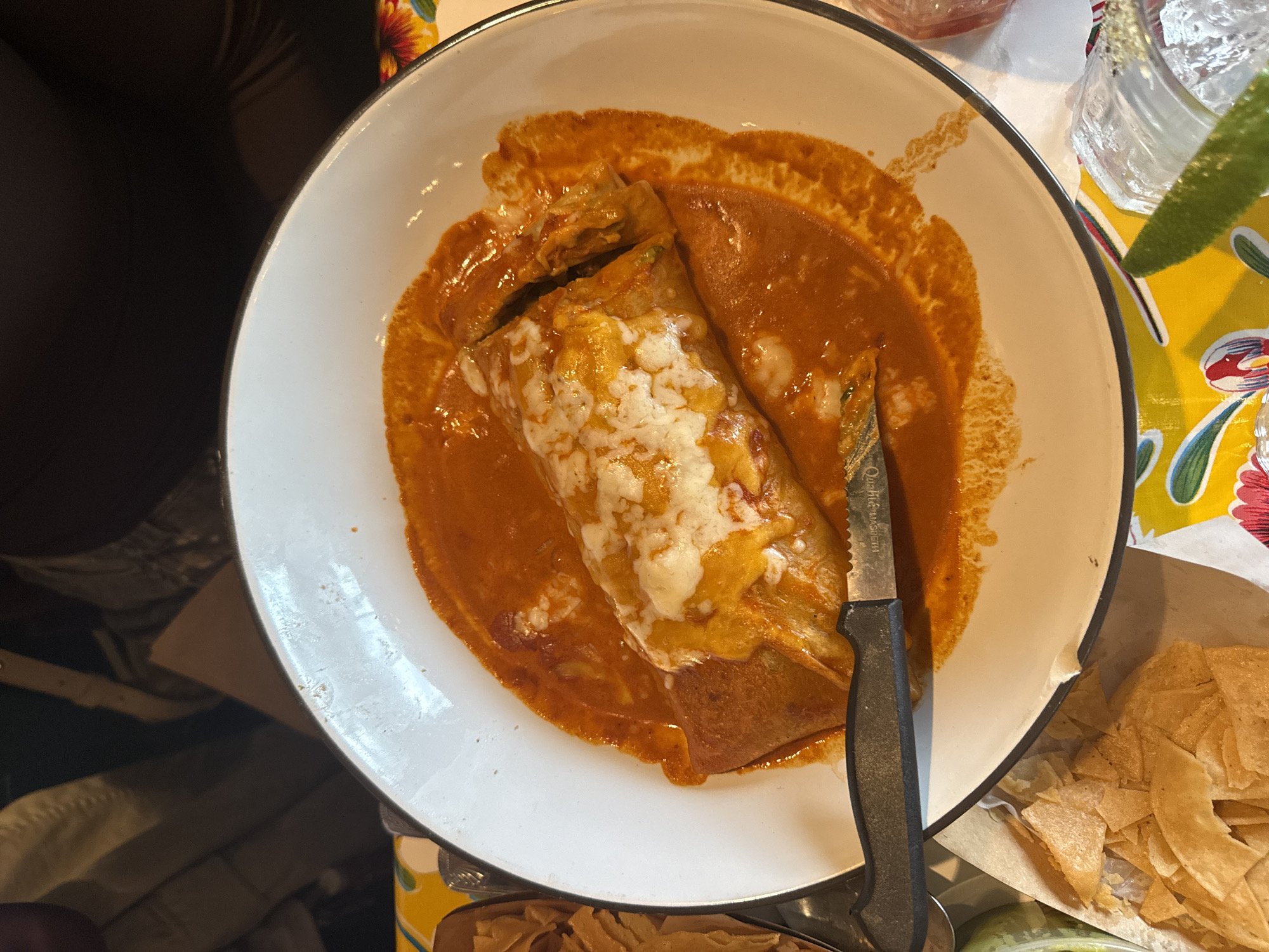 A close-up, high-angle shot shows a white bowl containing a burrito smothered in red sauce and topped with melted cheese. A black-handled knife partially rests on the burrito. To the right of the bowl, a glass with a lime wedge sits on a colorful tablecloth. A container of tortilla chips is also visible. The setting appears to be a restaurant in Los Angeles, United States of America. The person in the image is eating the burrito.