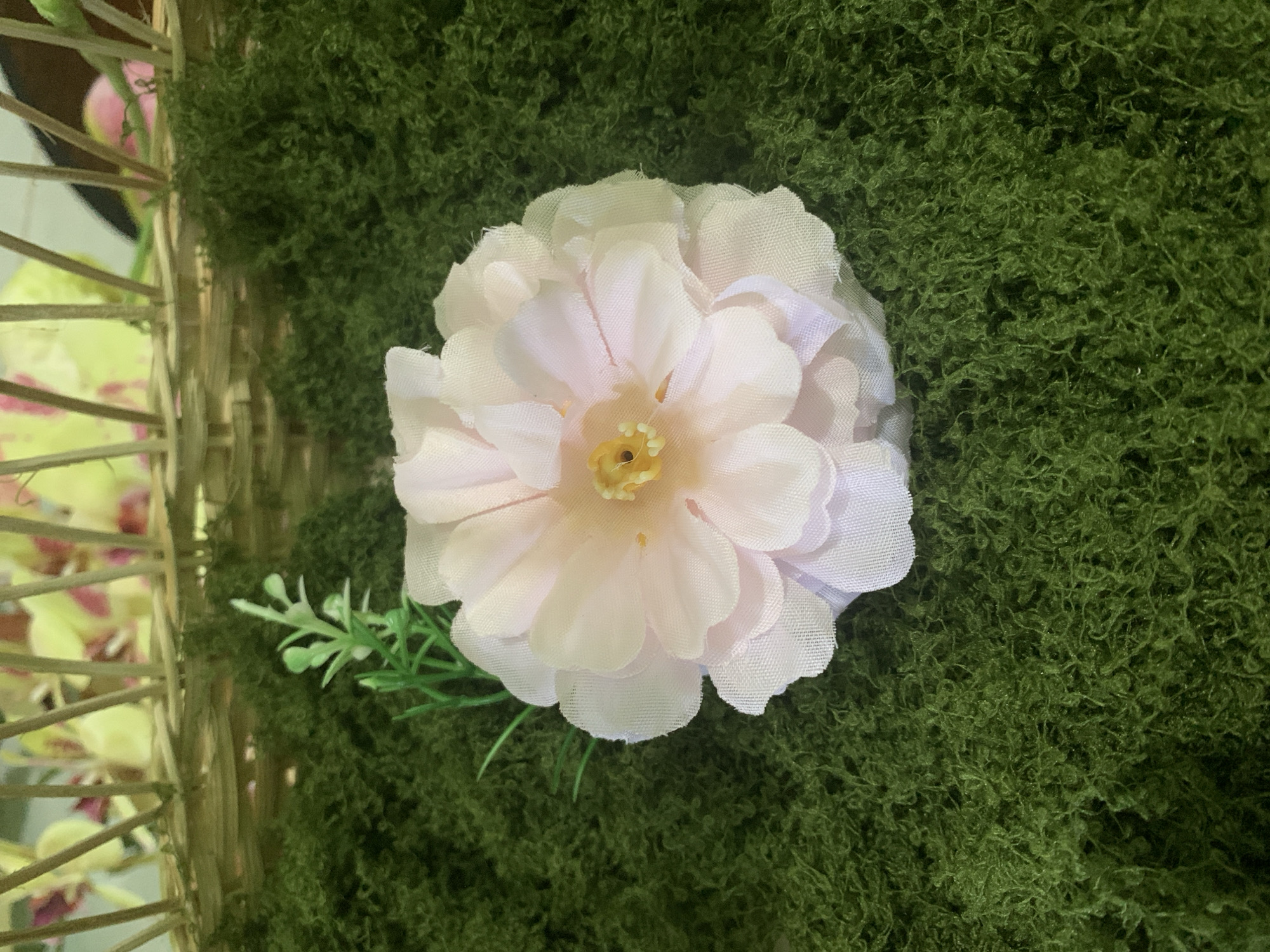 A single, pale pink artificial flower with delicate, layered petals and a yellow center is positioned on a bed of vibrant green artificial moss. To the left of the flower, a small sprig of artificial green leaves emerges from the moss. In the background, a portion of a woven basket is visible, containing blurred yellow and pink artificial flowers. The scene is illuminated with artificial light, casting subtle shadows. There are no people or animals present in the image. The image does not provide any information about its location in Kuala Pembuang, Indonesia.