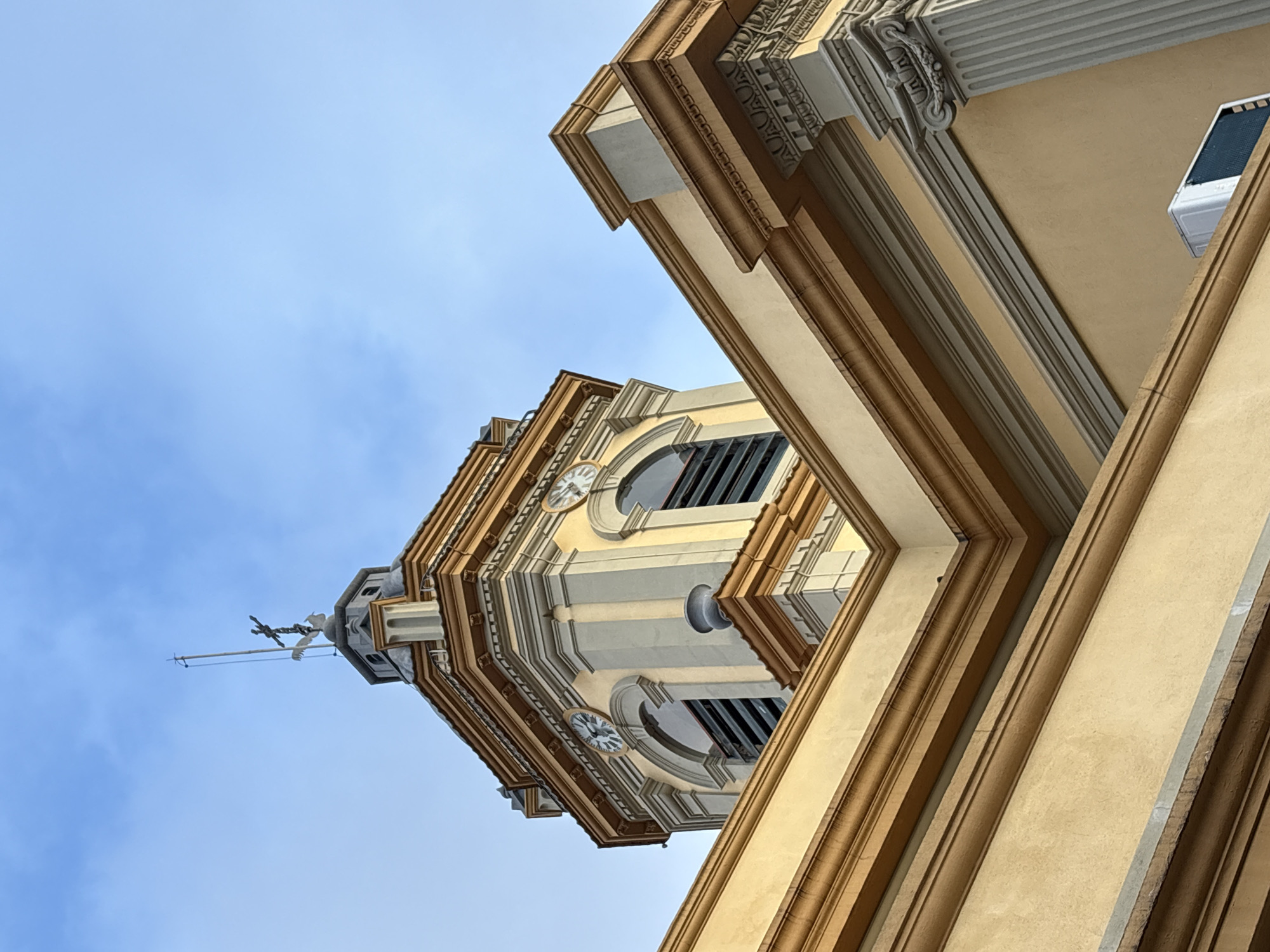 The image shows a low-angle view of the clock tower of the Igreja da Ordem Terceira de São Francisco, located in São Paulo, Brazil. The tower is painted in a light yellow color with contrasting brown and gray architectural details. Two clock faces are visible on the octagonal section of the tower, each showing approximately 11:30. At the very top of the tower, a cross with two rooster figures is mounted on a flagpole. The surrounding building, also in yellow, features decorative cornices and a fluted column. The sky above is blue with scattered clouds. An air conditioning unit is visible on the wall of the lower building. The scene is static, with no people or active events depicted.