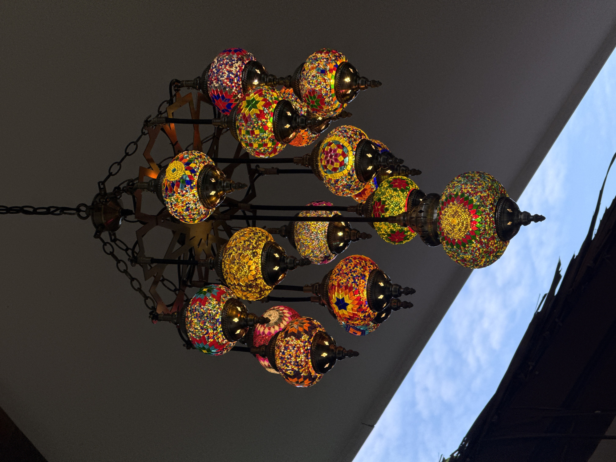 A chandelier with multiple glass lanterns is suspended from a dark ceiling. The lanterns are spherical and decorated with colorful mosaic patterns in shades of red, blue, yellow, green, and orange. Each lantern has a metal cap at the top and a pointed finial at the bottom, and lights inside illuminate the glass. The chandelier has a tiered structure with chains connecting the various lights.
The setting appears to be outdoors, as a glimpse of a blue sky with white clouds is visible in the upper right corner. The lighting suggests it is daytime. The overall impression is one of decorative illumination. No people are visible, and no text is present. The image captures a detailed view of the intricate craftsmanship of the mosaic lanterns.