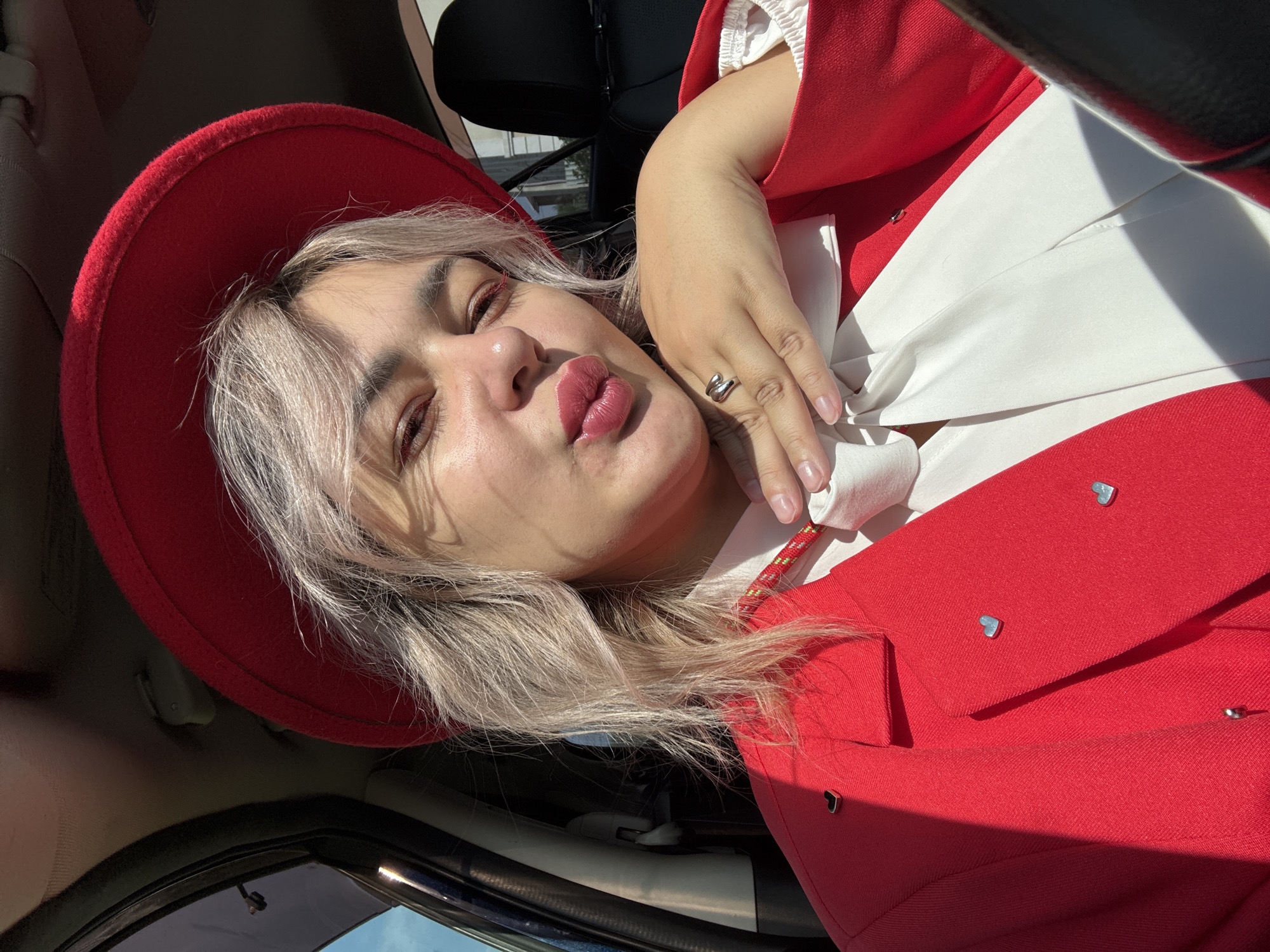 The image shows a close-up, waist-up view of a woman inside a car. She is wearing a red hat and a red blazer adorned with small silver heart-shaped pins. Beneath the blazer, she has on a white blouse with a large white bow. Her hand, adorned with a silver ring, is resting on her chin, and her lips are pursed. The lighting suggests it is daytime, with sunlight casting shadows across the scene. The background shows the interior of the car, including a dark seat and the car's ceiling. There is no visible indication of the city of Birobidzhan, Russia, or any specific urban environment.
