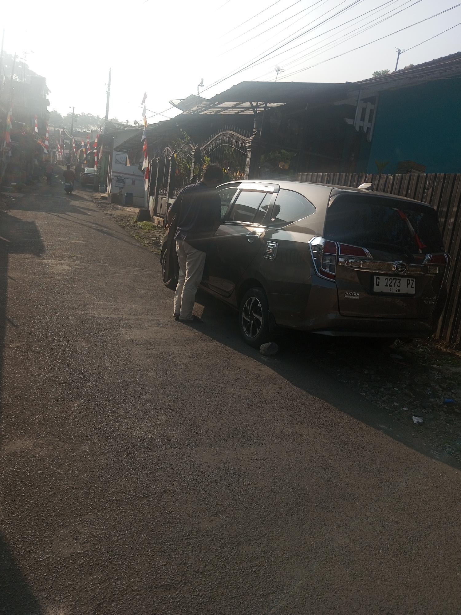 In Krasak, Indonesia, a man in a dark shirt and light-colored pants stands beside the open driver's side door of a dark brown hatchback car. The car is parked on an asphalt road, which curves slightly to the left. The car's license plate reads "G 1273 PZ". To the right of the car, a wooden fence runs along the side of the road. In the background, several houses with tin roofs are visible, and a street is lined with Indonesian flags. The scene is illuminated by bright sunlight, casting long shadows across the road.