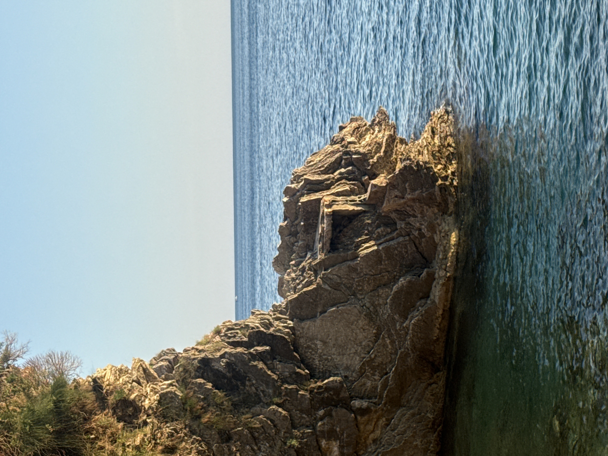 The image depicts a rocky outcrop on a coastline, with the deep blue sea stretching out to the horizon. The sun appears to be high in the sky, casting strong shadows on the rugged terrain and highlighting the texture of the rocks. The water's surface is rippled, indicating a gentle breeze or slight chop. The scene is natural and unpopulated, with no people or man-made objects visible. The vegetation on the rocky slopes is sparse, with some hardy shrubs and trees clinging to the crevices. The overall mood is one of natural beauty and tranquility, with a sense of raw, untamed wilderness. The image appears to be taken at eye level, with the camera pointed slightly downwards towards the sea. The lighting and composition suggest a clear, sunny day, likely during the warmer months. The rocky outcrop is the central focus, with its layered structure and weathered appearance conveying a sense of geological history. The juxtaposition of the solid, ancient rock with the fluid, ever-changing sea creates a dynamic and visually engaging composition.