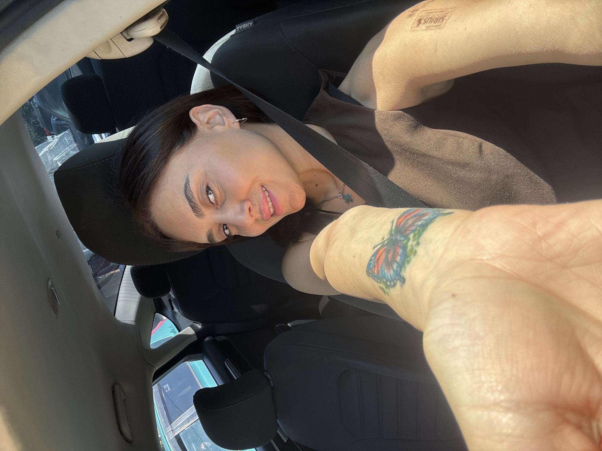 In the image, a woman is seated in the driver's seat of a car. She has dark hair and is wearing a dark top and a seatbelt. A butterfly tattoo is visible on her left arm. The interior of the car, including the dashboard and seats, is visible. The sunlight suggests it is daytime. There is no visible indication of the location being in Tbilisi, Georgia.