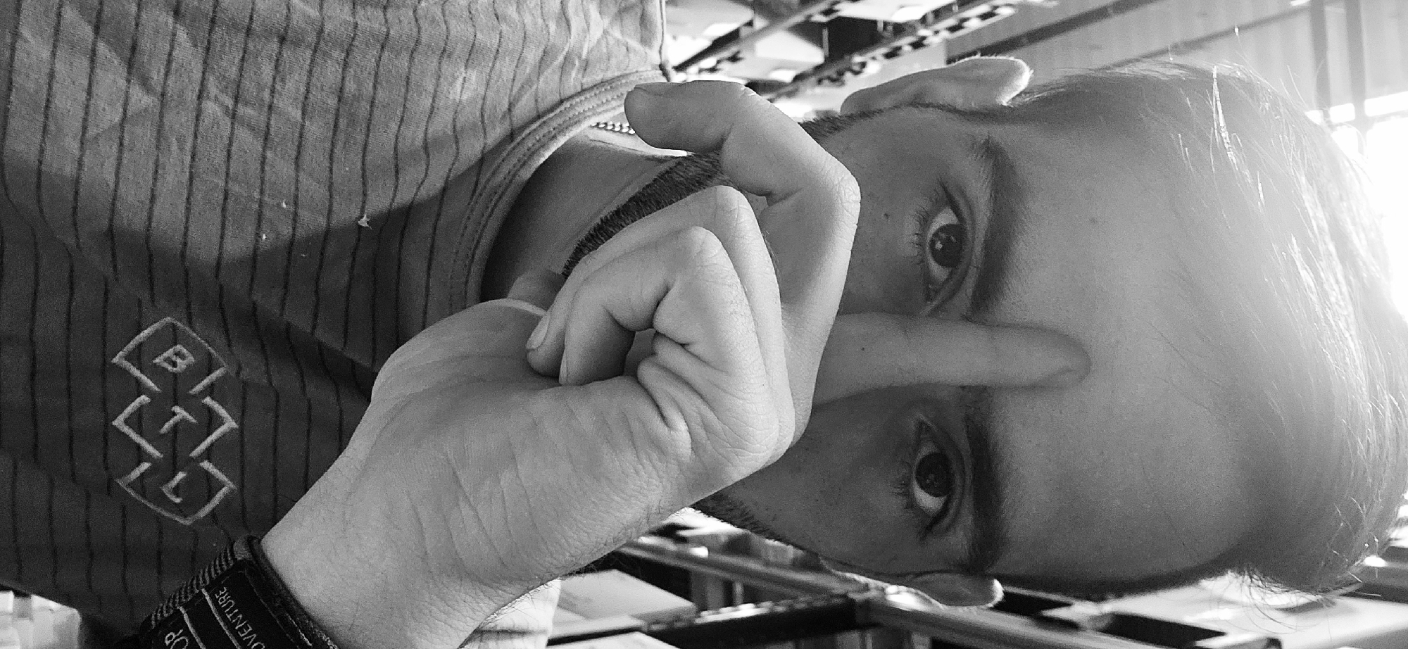 A medium-shot black and white image shows a man's face and hand in the foreground, oriented as though the viewer is looking up at him. The man's head is tilted to the left; his eyes are visible, and the bridge of his nose is covered by a finger of his left hand, which is clenched into a fist. He has short hair, and a stubble beard is faintly visible. Part of his upper torso is visible, wearing a vertically striped t-shirt with a logo that reads "BTL." A watch with the word "ADVENTURE" on the wristband is on his left wrist. The background shows a blurred environment with metallic structures and bright sunlight shining through a window or opening. It is not possible to determine the specific location in Plovdiv, Bulgaria, from the visual information provided.