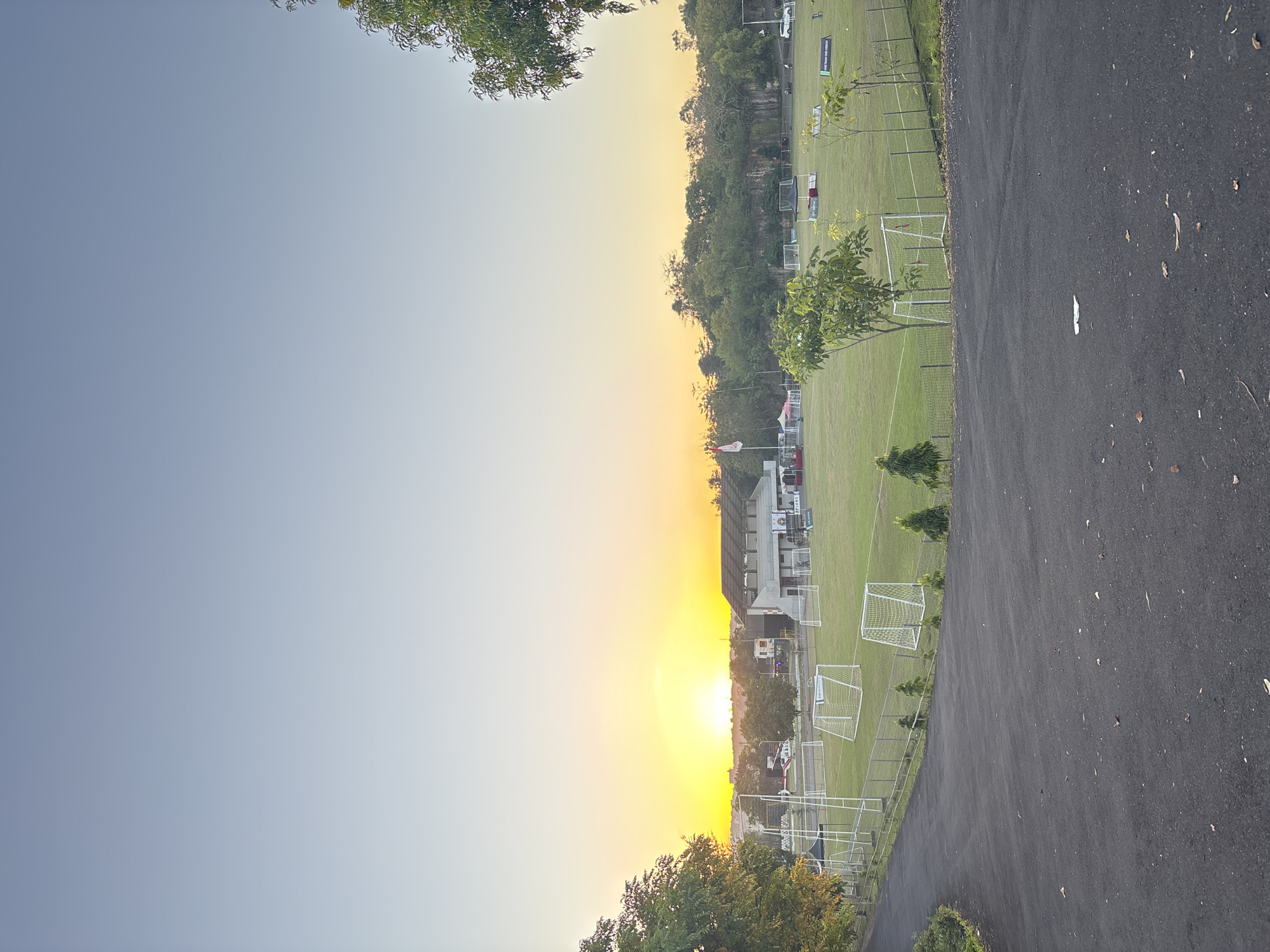 The image depicts a soccer field at sunset, with a parking lot adjacent to the field. A building is located near the field, and trees border the area. The location is Kuta Selatan, Indonesia, based on the presence of soccer goals which are common in the area. There are no people visible in the scene.