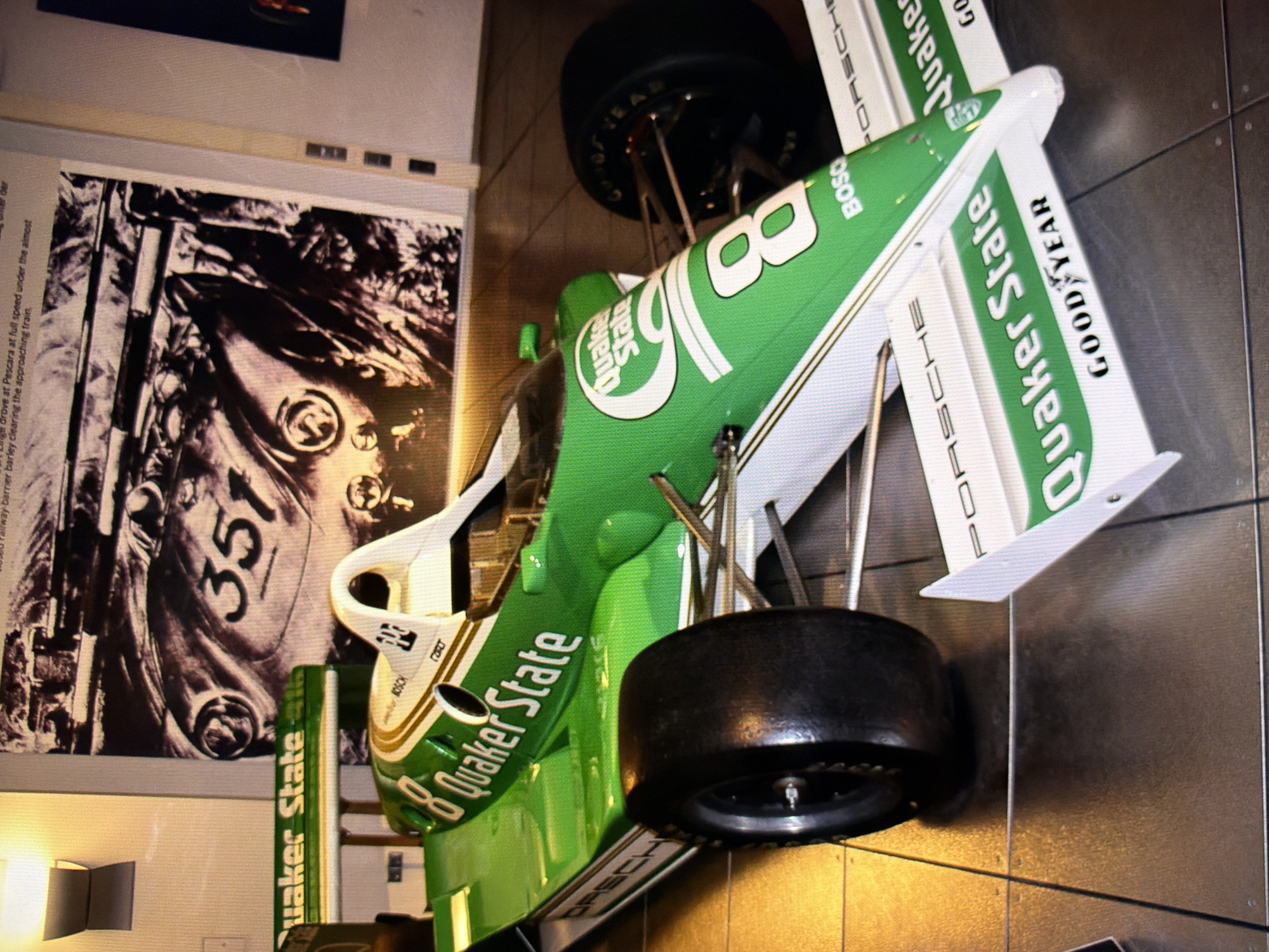 The image shows a green and white race car with "Quaker State" and "Goodyear" branding. The car is on display indoors, likely in a museum or exhibition space. The background features a large black and white photograph of another race car with the number "351". The flooring appears to be tiled. The image does not provide any information about the location being Kyiv, Ukraine.
