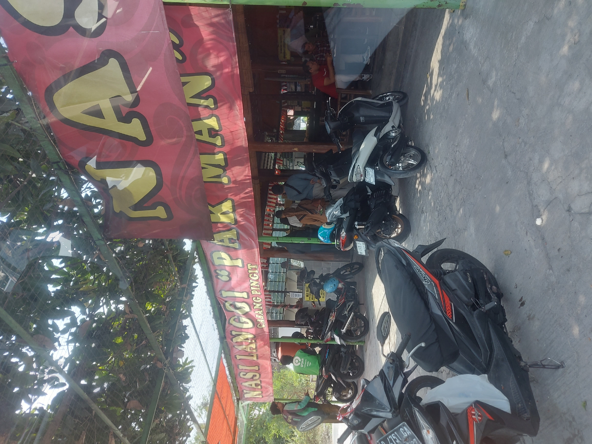 The image depicts an outdoor dining area at a restaurant called "NASI LANGGI 'PAK MAN' CABANG PINGIT" in Kricak, Indonesia. A large red banner with yellow lettering advertising the restaurant's name is prominently displayed. Several motorcycles are parked in front of the establishment, suggesting a popular spot for local transport. Some individuals are seen entering or exiting the restaurant, while others are seated inside. The environment appears to be a casual eatery with a covered seating area and a concrete floor. Lush green foliage hangs from above, providing shade and a natural ambiance.