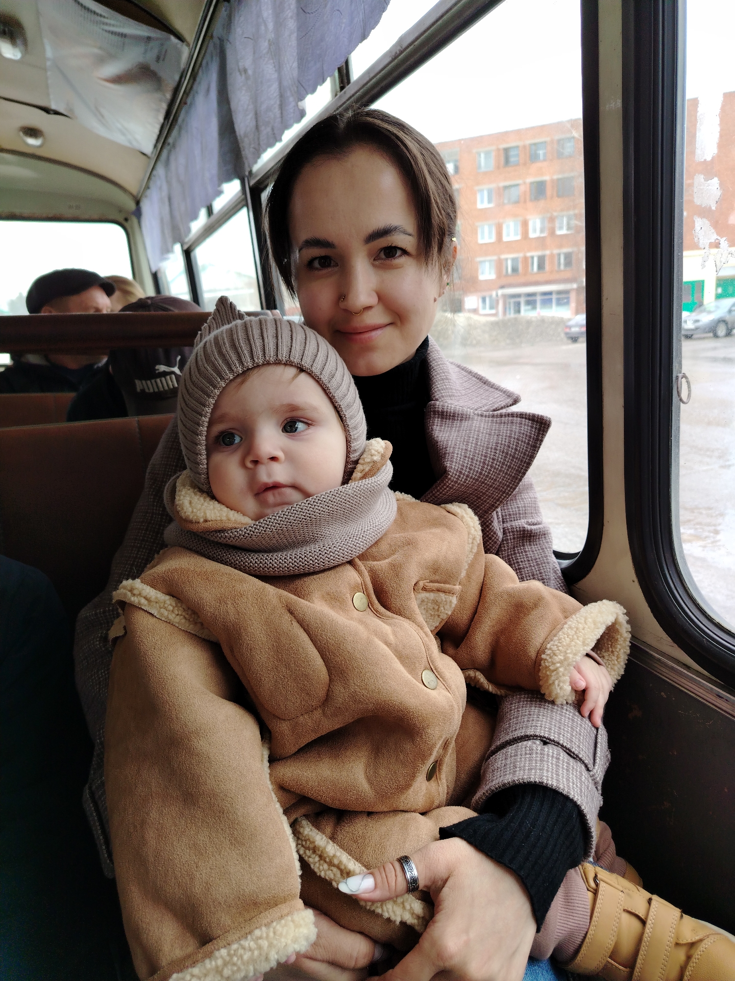 A woman and a baby are seated inside a bus or tram. The baby is wearing a brown jacket with a fur-like lining and a matching knit hat and scarf. The woman has dark hair pulled back, a nose ring, and is wearing a brown patterned coat over a black turtleneck. The bus's interior has brown seats and white curtains above the windows. Outside the window, a multi-story brick building is visible. The location is likely in Noviy, Russia, based on the style of the building and bus design.