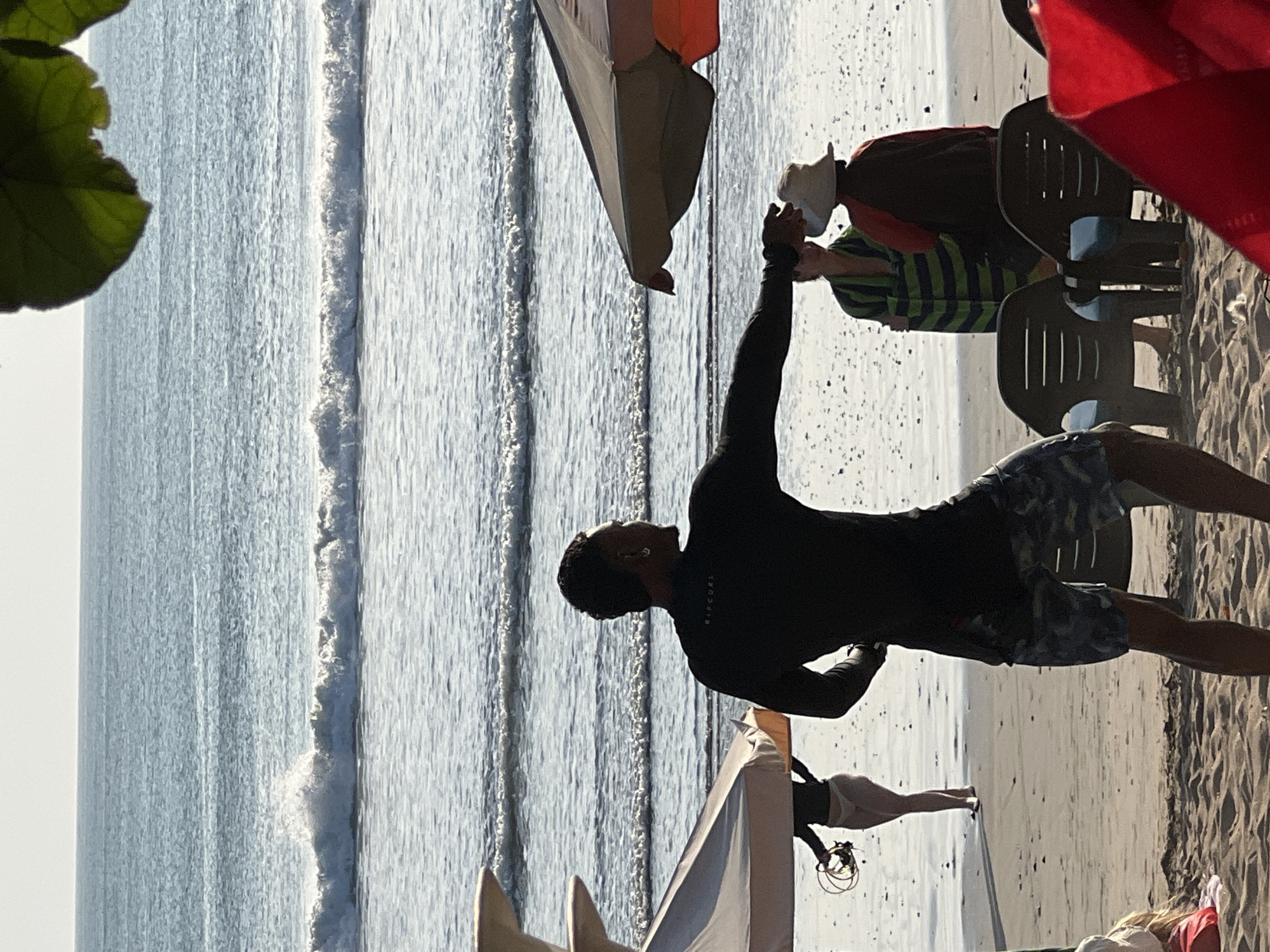 At Kuta Beach, Bali, Indonesia, under a bright, clear sky, the ocean exhibits gentle waves. In the foreground, a man wearing a black rash guard and patterned shorts stands facing away from the camera, with his right arm extended. To his left, a woman in white bikini bottoms and a black long-sleeved top stands near an umbrella, holding a round object. In the middle ground, two individuals are conversing, one wearing a green and black striped towel or wrap, and the other a sun hat and a rust-colored top. Several plastic chairs are arranged on the sand. A large, colorful umbrella is partially visible on the right side of the frame. The sand appears wet and textured, with small pebbles and debris scattered across it.