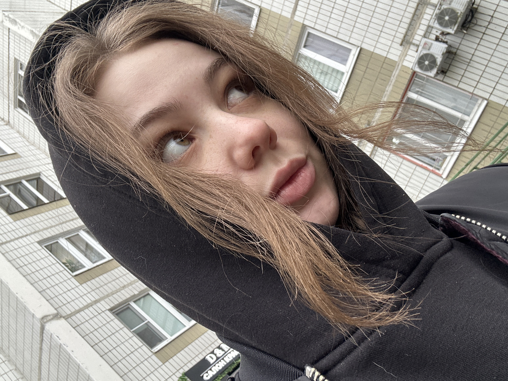 A young woman is angled upward, wearing a black hoodie with the hood up. Her light brown hair is partly covering her face. The background consists of a multi-story building, suggesting a city setting. A sign that reads "D&A САЛОН КРА" is partially visible. The building's facade is light-colored, with rows of white-framed windows and light brown accents. Two air conditioning units are visible on the building. Based on the Cyrillic text on the sign, the location is likely Moscow, Russia.