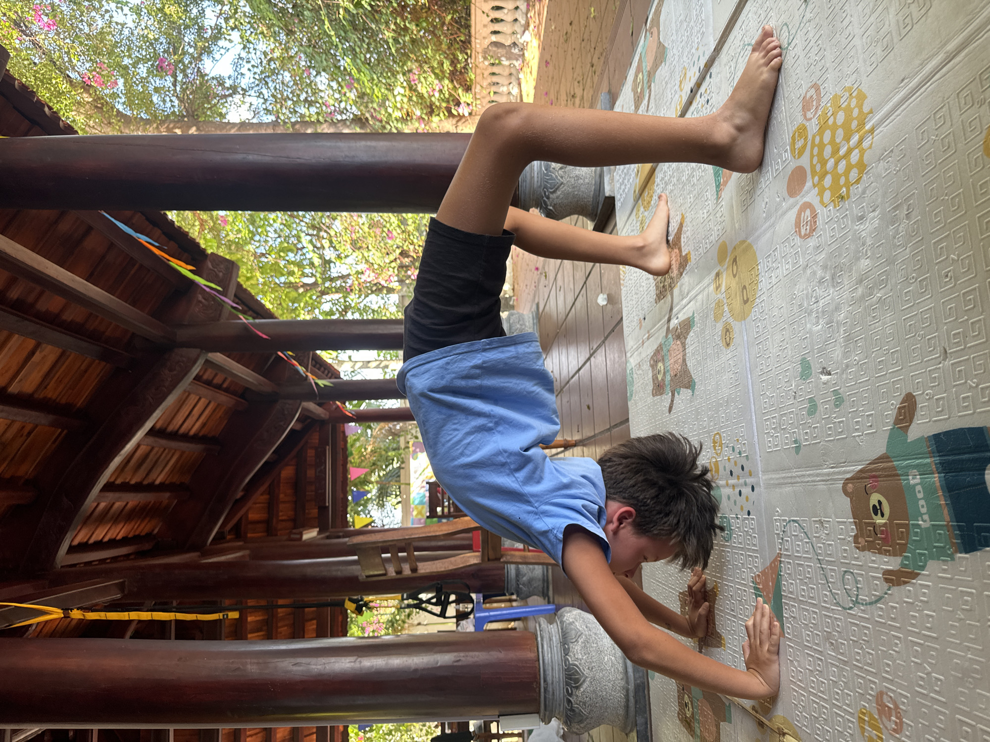 A child is performing a handstand indoors on a white mat with cartoon bear designs, in Mũi Né, Vietnam. The child, wearing a blue t-shirt and black shorts, is positioned upside down with their feet against a wall, hands on the mat, and body forming a straight line. The background includes a wooden structure with pillars and a roof, suggesting the interior of a building or outdoor covered area. There are colorful flags hanging and foliage visible through the wooden structure.