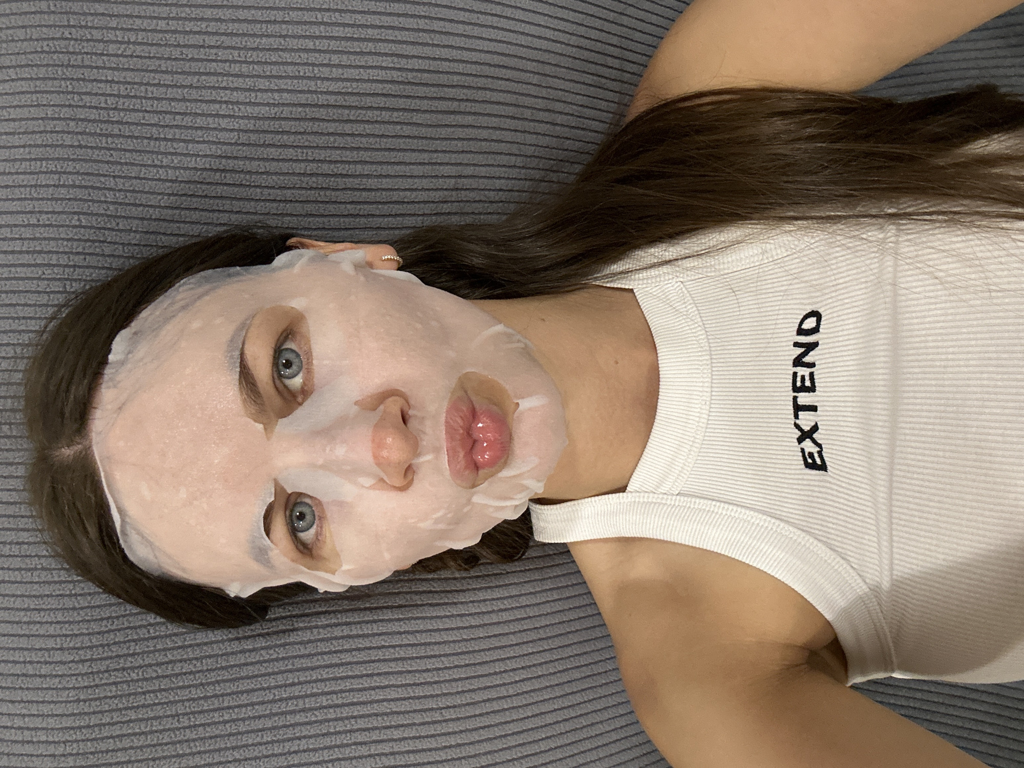 A woman is lying on a gray textured surface, likely a bed or couch. She is wearing a white tank top with the word "EXTEND" printed on it. A sheet mask covers her face, with openings for her eyes and mouth. Her hair is long and brown. The background is the same gray textured surface. The image does not contain any information indicating the location in Budva, Montenegro.