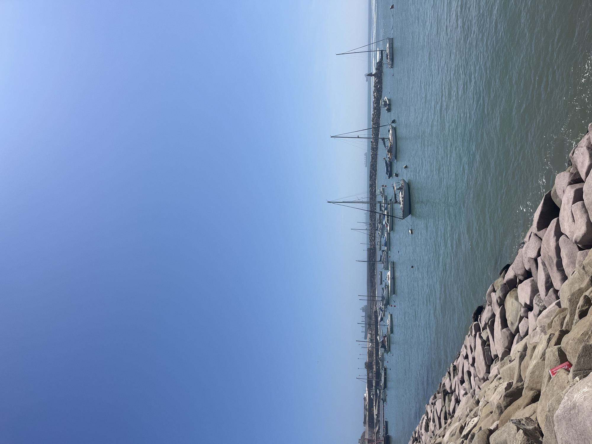 The image depicts a harbor scene in Arica, Chile, taken from a position on a rocky breakwater. The breakwater consists of large, angular stones. To the left of the breakwater, the harbor is filled with numerous sailboats moored along a dock, their masts reaching vertically. The water is a muted blue-green. A city skyline is visible in the background, beyond the boats. The sky is a clear, gradient blue, lighter near the horizon and darker above. A faint haze is present in the distance, affecting the visibility of the horizon line. A discarded red-colored rectangular object rests on the rocks of the breakwater. No people are visible in the image.