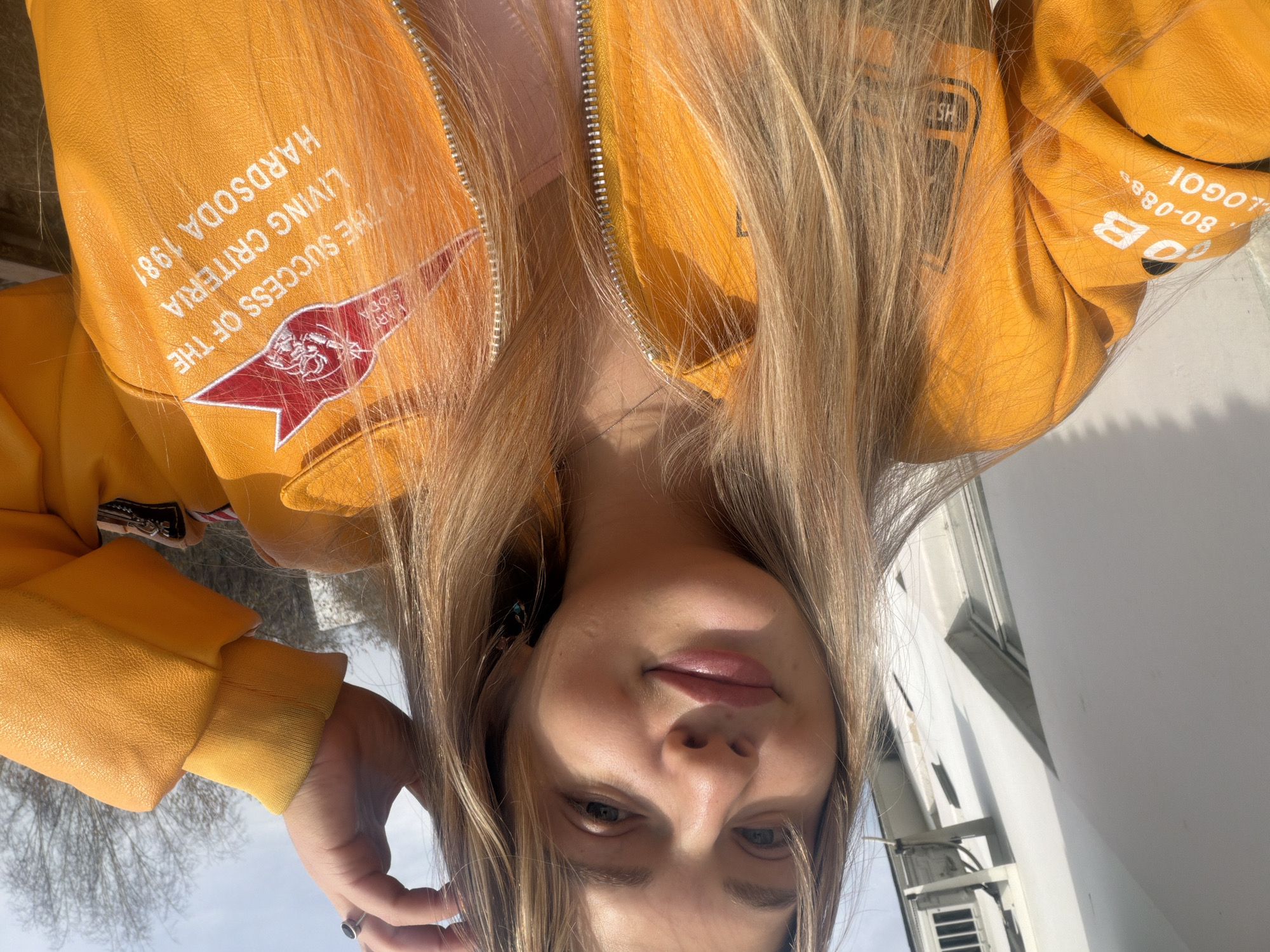 A woman is posing in an upside-down selfie in an outdoor location in Naberezhnye Chelny, Russia. The woman has blonde hair and is wearing a bright yellow jacket with patches that read "The Success of the Living Criteria, Hard Soda 1981" and "Logo 80-0888 OB SH". She appears to be standing in front of a white building with a window air conditioning unit. The sky is light blue, and some bare trees are visible in the distance.