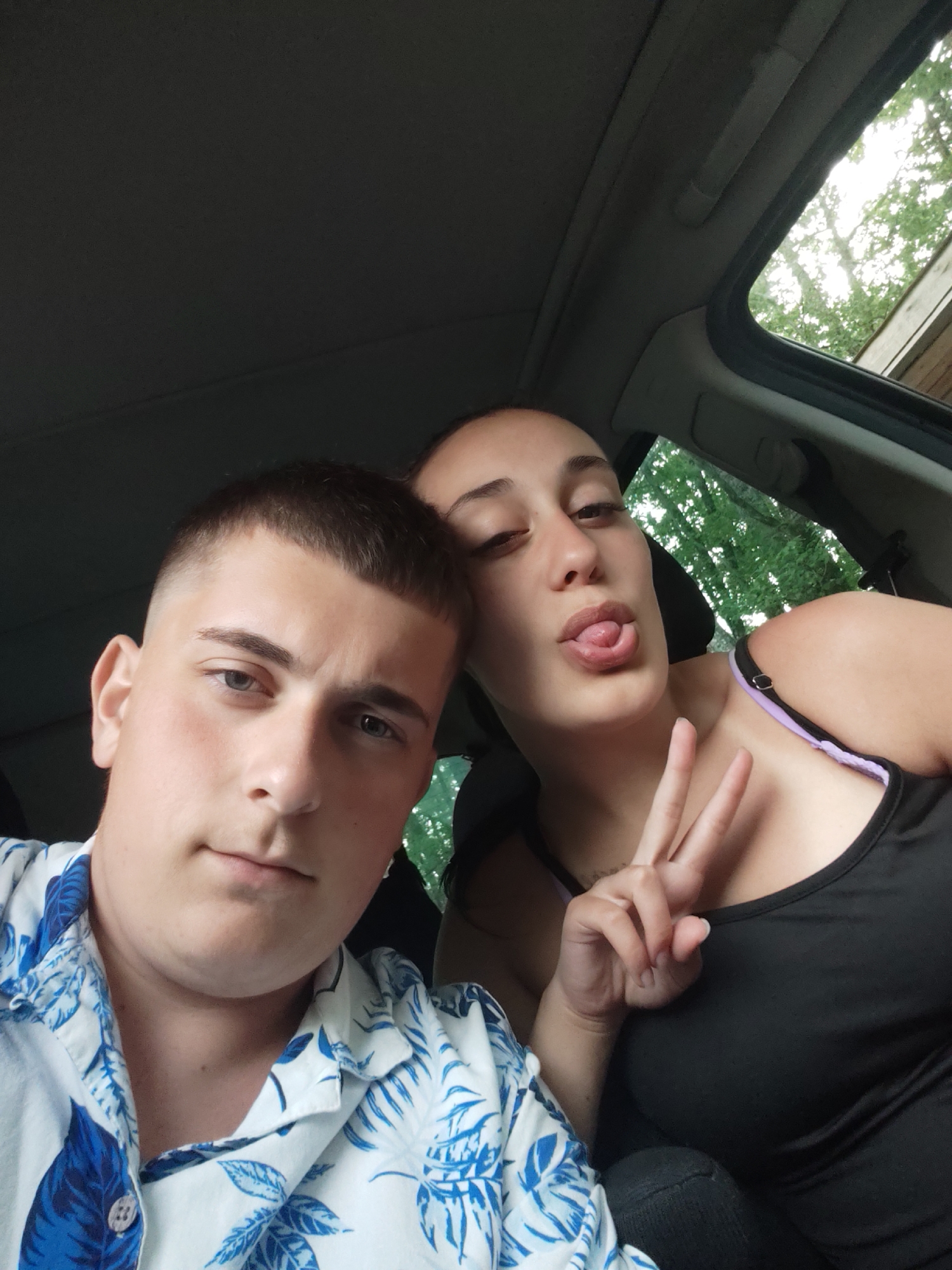 This selfie captures two young individuals inside a vehicle, likely during the daytime in Maglishta, Bulgaria.
On the left, a young man with short, dark hair and light eyes gazes directly at the camera with a neutral expression. He is wearing a light-colored, short-sleeved button-up shirt featuring a distinctive blue tropical leaf pattern. To his right and slightly behind him, a young woman is visible, making a playful gesture by sticking her tongue out and holding up a peace sign with her right hand. She wears a dark tank top with a visible purple strap.
The car's interior, including the headliner and parts of the window frames, is visible. Through the side windows and sunroof, bright green foliage and a clear sky can be seen, indicating it is daytime. The overall scene suggests a casual, perhaps travel-related, moment between the two subjects. No text is visible in the image.