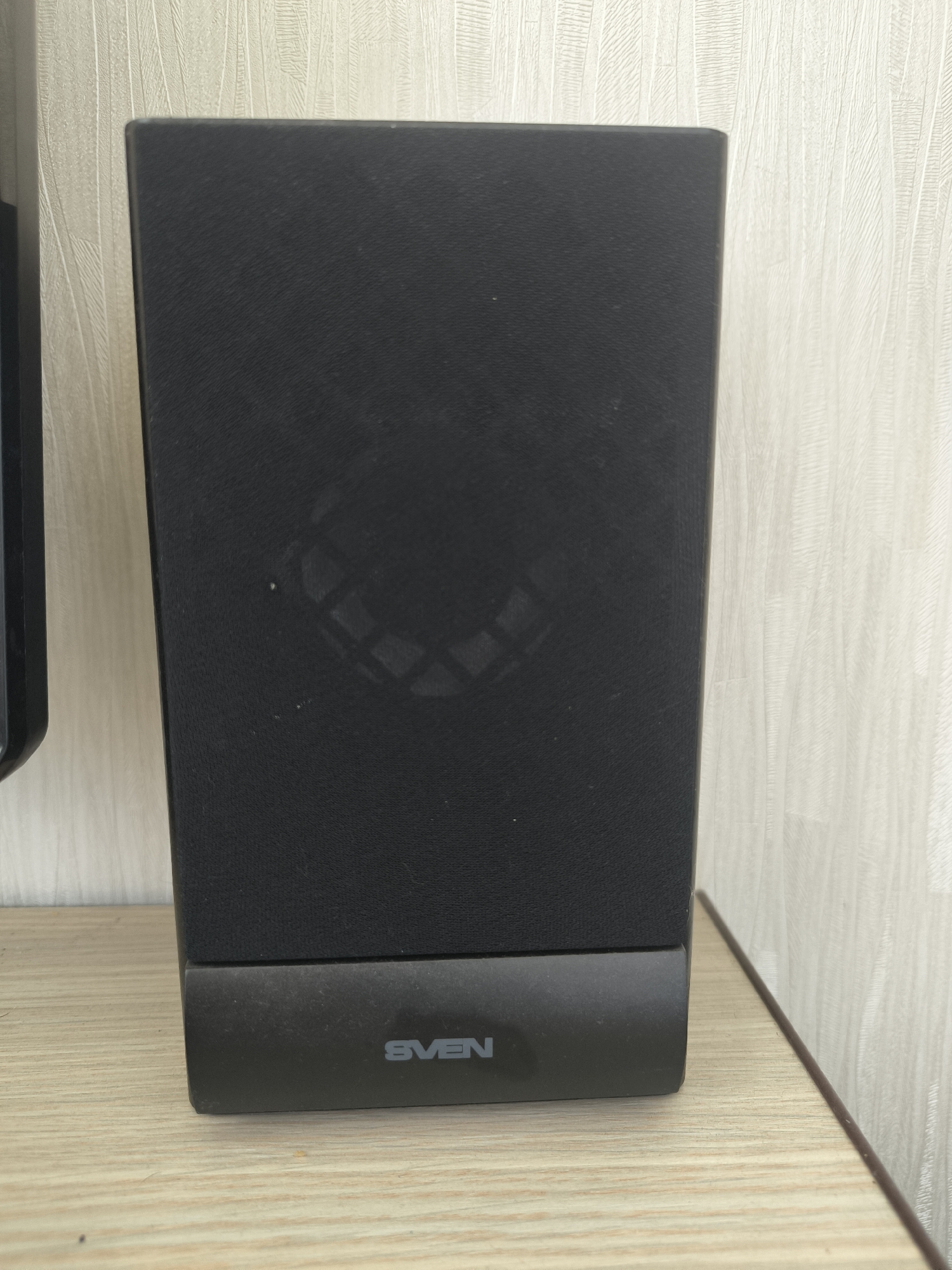 A black speaker with the brand name "SVEN" visible in silver lettering on its base sits on a light wood-grain surface. The speaker is positioned in front of a textured beige wall. There are no people or actions depicted in the image, and no specific location in Chișinău, Moldova can be determined from the visual information. The scene is static and focuses solely on the speaker and its immediate surroundings.