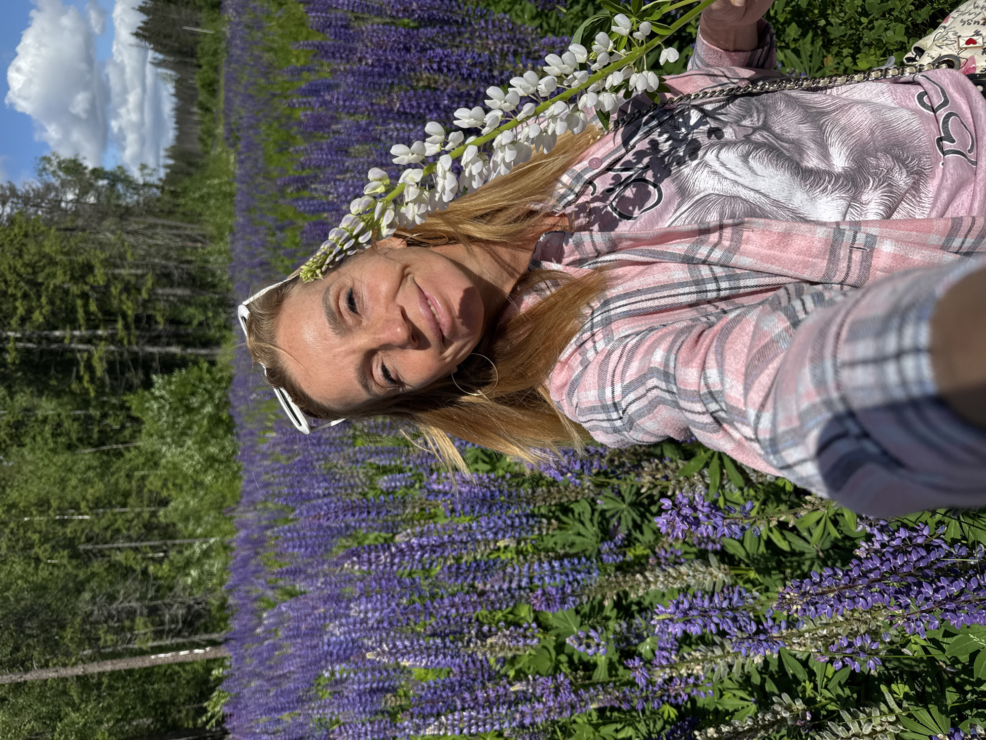 A woman with blonde hair, wearing white-rimmed sunglasses on her head, a pink plaid shirt over a graphic t-shirt, and large hoop earrings, poses for a selfie in a field of purple lupine flowers. A stalk of white lupine flowers is held near her face. Behind her, a dense forest of green trees is visible under a bright blue sky with scattered clouds. The scene appears to be in a rural or natural setting, possibly during daytime. The image is a close-up shot of the woman, with the flowers and forest serving as the backdrop.
