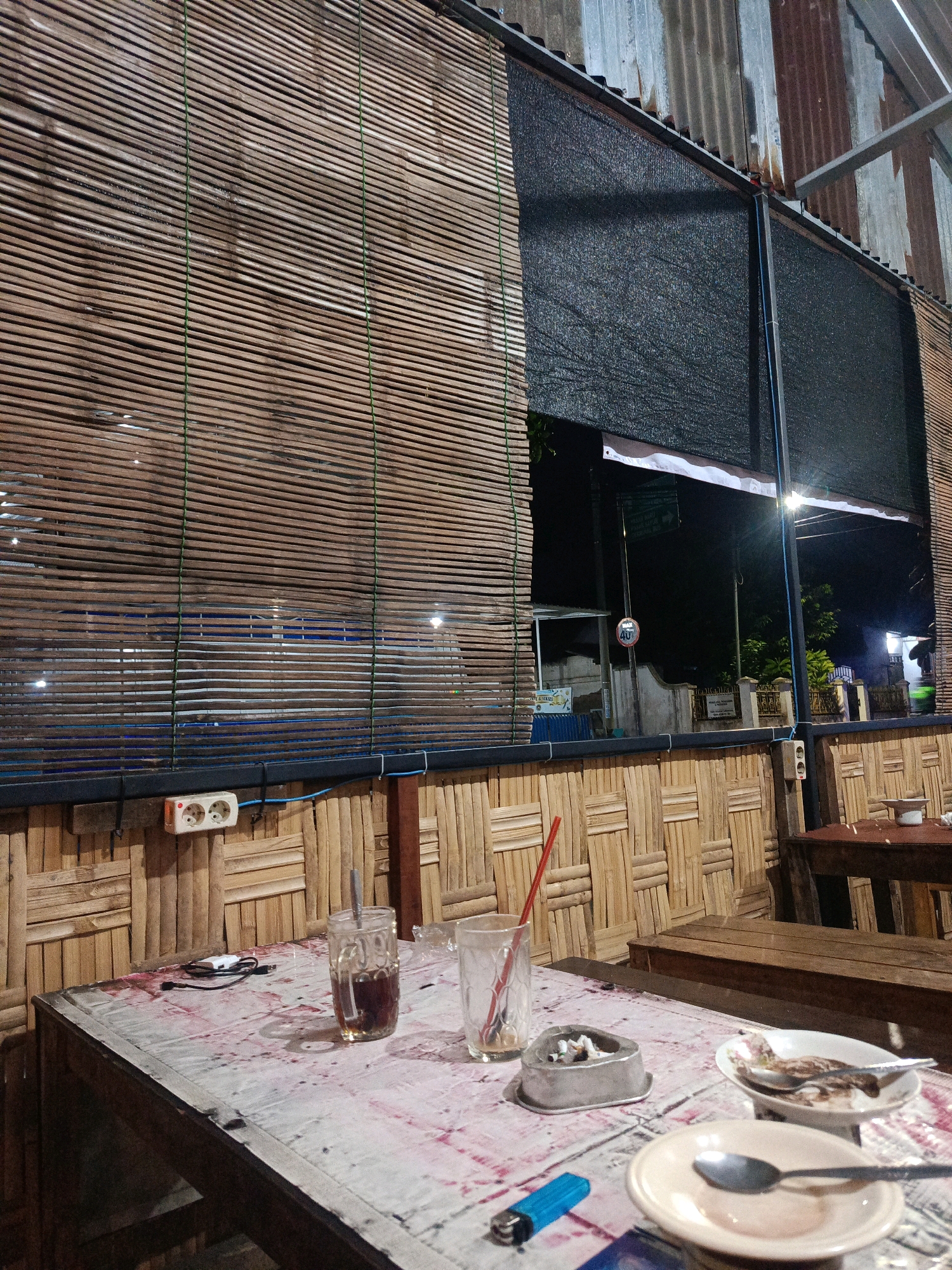 The image depicts an outdoor seating area, likely a cafe or warung, in Magetan, Indonesia. The setting is rustic, with walls made of woven bamboo and a roof constructed from corrugated metal. A bamboo blind covers a large portion of the wall.
On a wooden table in the foreground, several objects are present, suggesting recent activity. These include two glasses, one with a red straw and dark liquid, and another empty glass with a red straw. An ashtray filled with cigarette butts and a blue lighter are also on the table. Two small plates with remnants of food and spoons are also visible. A USB cable and a small white object are also on the table.
The background shows a glimpse of the outside environment, including a street with signs and other structures, illuminated by artificial lighting. The overall impression is of a casual, open-air establishment at night.