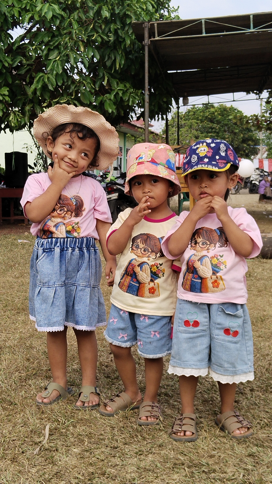 In Margatunggal, Indonesia, three young girls stand outdoors on a grassy area under a canopy. The girl on the left wears a straw hat, a pink t-shirt with a graphic of a girl in glasses and holding flowers, and a blue denim skirt with white trim. She rests her chin on her hand and smiles. The middle girl wears a pink bucket hat, a cream-colored t-shirt with a similar graphic to the first girl, and blue denim shorts with pink bow prints and white trim. She has her fingers to her mouth. The girl on the right wears a blue baseball cap with a yellow brim and cartoon graphics, a pink t-shirt with the same graphic as the other two girls, and blue denim shorts with cherry prints and white trim. She also has her fingers to her mouth. Behind the girls, trees and a structure with a brown tarp roof are visible.