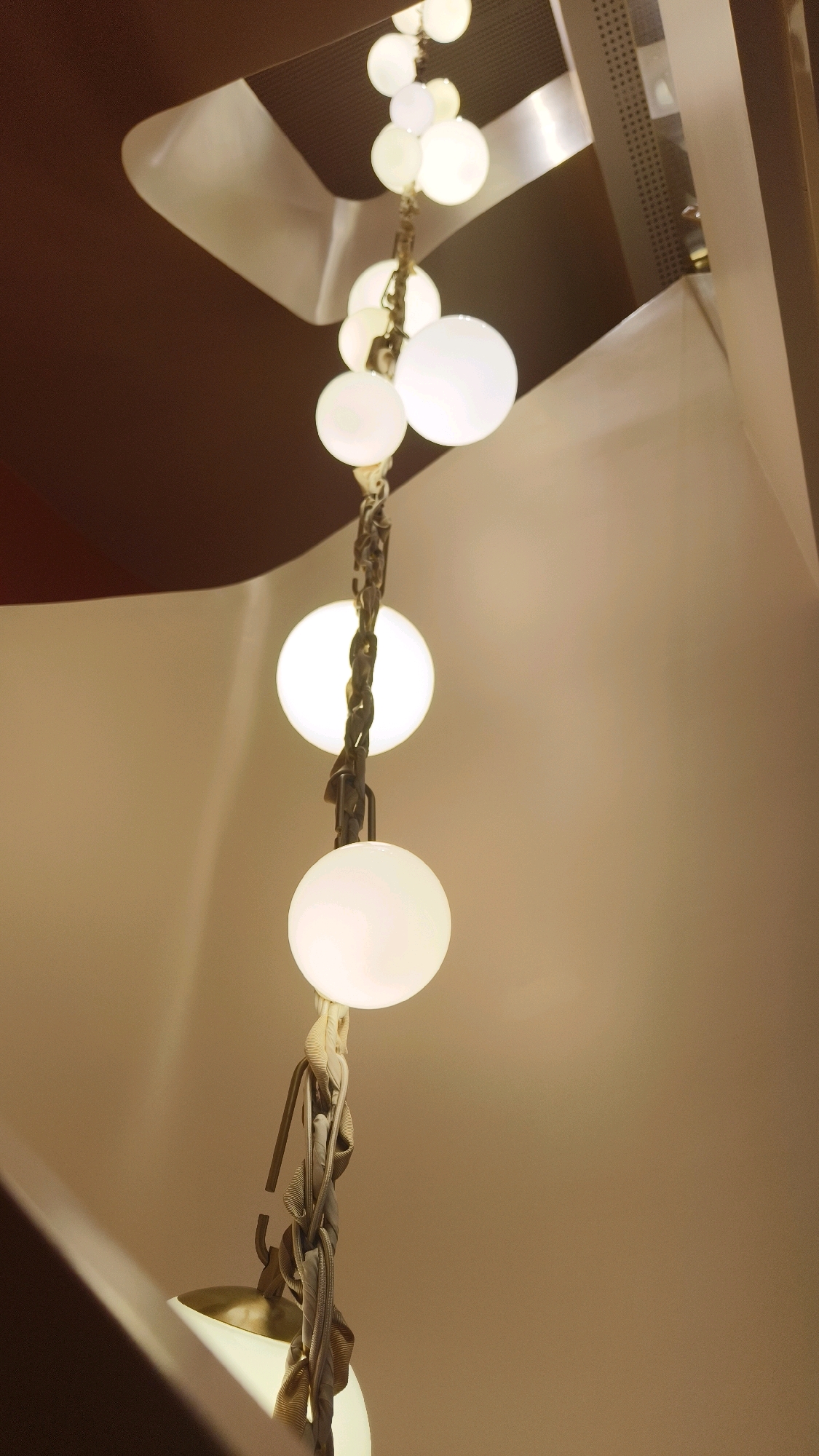 The image depicts a close-up, angled view of a modern chandelier with several spherical, frosted glass lights suspended by a decorative chain. The lights are illuminated, casting a warm glow. The chandelier is hanging against a backdrop of neutral-colored walls and ceiling structures, possibly within a building's interior, such as a staircase or foyer. The lighting suggests an indoor setting, with the warm tones of the light and the surrounding surfaces indicating an ambient atmosphere. There are no people visible in the image, nor any indication of a specific location within Vienna, Austria. The focus is entirely on the design and illumination of the chandelier.