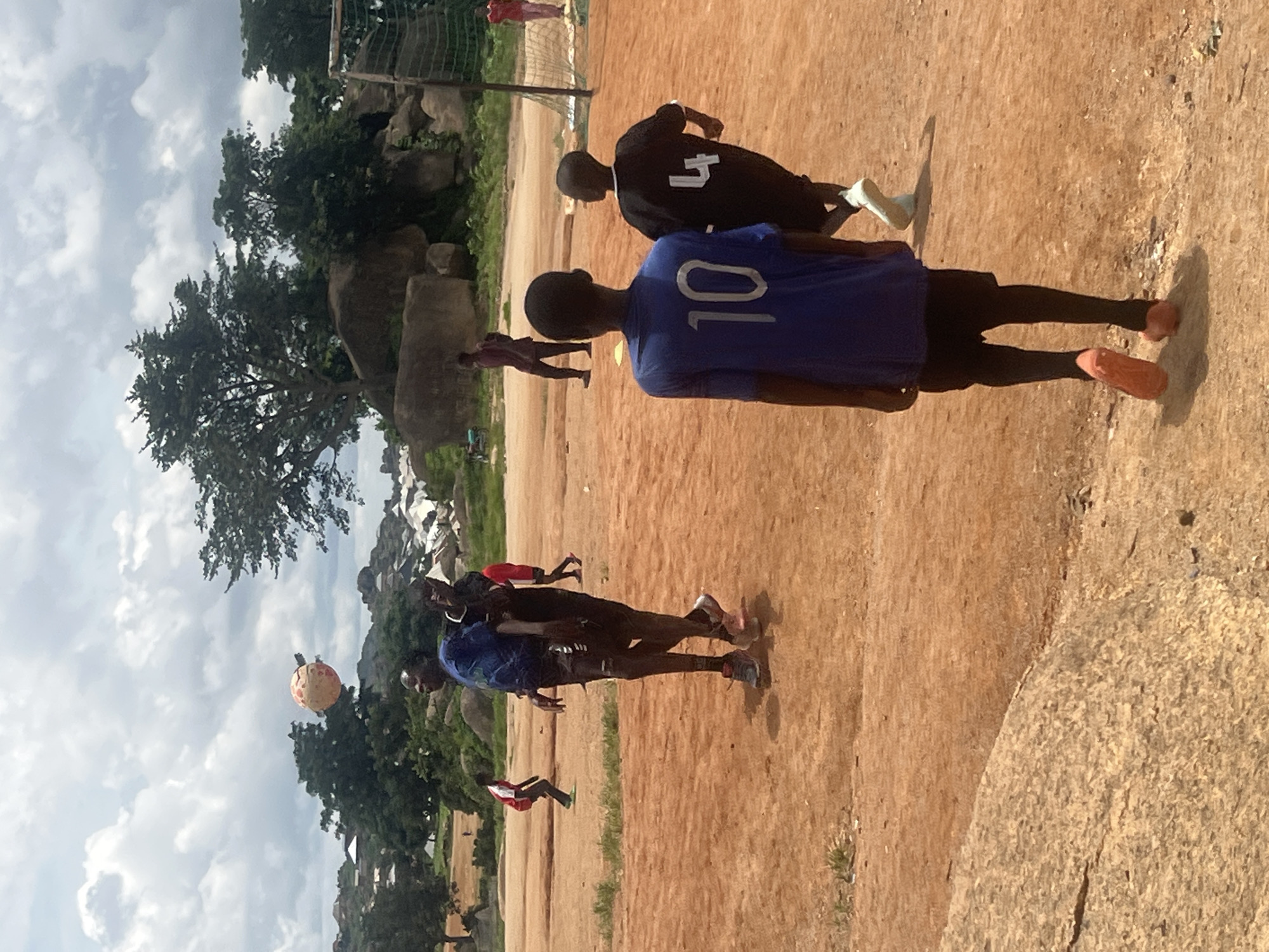 A group of boys are playing soccer on a dusty field in Jos, Nigeria. The sky is overcast with scattered clouds. In the foreground, two boys are walking away from the camera, one wearing a blue shirt with the number "10" and black shorts, the other in a black shirt with the number "4" and black shorts. In the midground, a group of boys are in action with a ball in the air. One boy in a blue shirt appears to be jumping to head the ball, while another boy in a black shirt is close by. Further in the background, other children and adults are visible, some watching the game and others walking around. The field is dirt and sparse with some patches of dry grass. Rocks and trees are scattered throughout the landscape, and in the distance, buildings and tents can be seen. A soccer net is set up on the right side of the field.