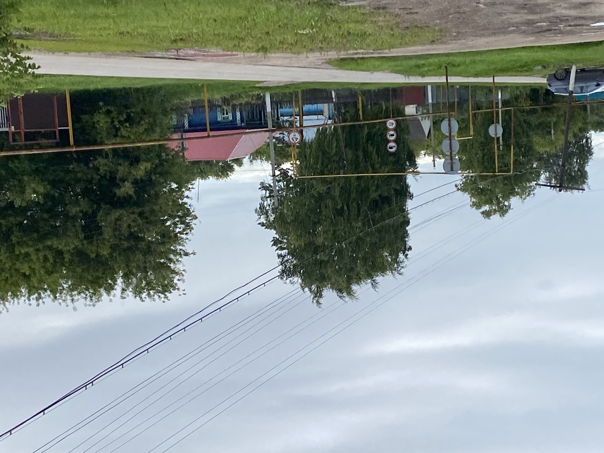 The image depicts a street scene in the village of Nadezhdino, Russia, under an overcast sky. In the foreground, a paved road curves to the right, with grassy verges on either side. A dark grey car is parked on the left side of the road.
Further into the scene, a yellow metal frame structure with a horizontal bar runs across the mid-ground. Attached to this structure are several traffic signs. On the left side of the frame, there are three circular signs stacked vertically. The bottom sign indicates a speed limit of "40". To the right of this, a traffic light with three circular lights is mounted on a yellow pole. Further to the right, another set of three circular traffic signs is visible, with the bottom one also displaying "40".
Behind the yellow frame, several houses are visible. A blue wooden house with a red roof stands prominently. To its left, a smaller white house with a dark roof is partially visible. A dark wooden fence runs along the front of the properties. Dense green trees and foliage are scattered throughout the background, with a very large, tall tree dominating the center-left of the image. Power lines stretch diagonally across the sky from the upper left to the upper right of the frame. The environment appears rural and somewhat weathered, with the overall impression being that of a quiet village street.