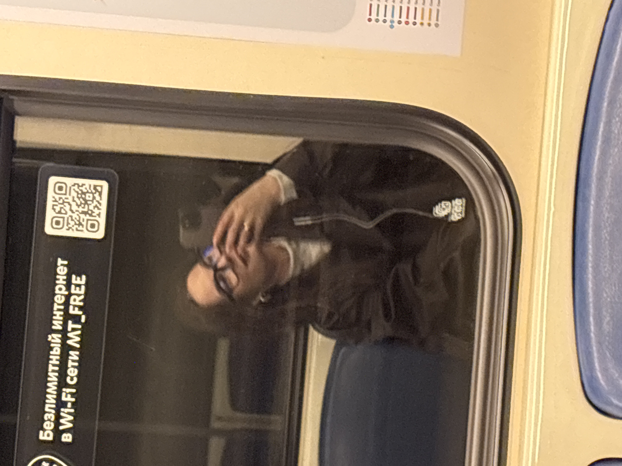 The image depicts the reflection of a person with glasses in the window of a Moscow Metro train. A sticker is affixed to the glass with a QR code and text in Russian, "Безлимитный интернет в Wi-Fi сети MT_FREE" (Unlimited internet in MT_FREE Wi-Fi network). The person in the reflection is wearing a dark jacket and appears to be seated. The train interior is visible, including a portion of a blue seat. Part of a route map with colored circles is visible above the window. The location is Moscow, Russia, specifically on a train in the Moscow Metro system.