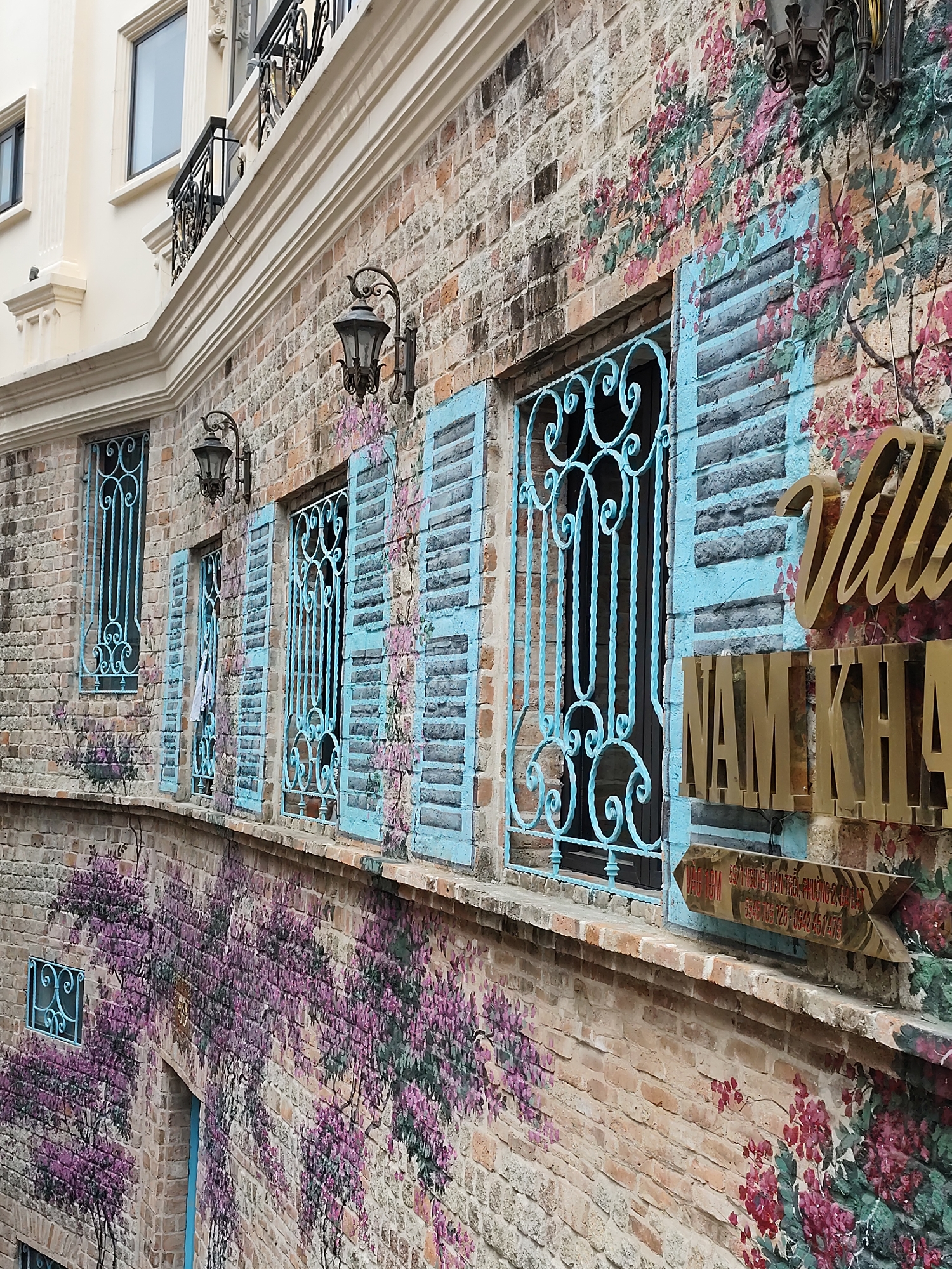 The image depicts the exterior of Villa Nam Kha in Da Lat, Vietnam. The building has a brick facade adorned with painted floral designs, including purple wisteria and pink blossoms. Windows are framed with blue ornamental wrought iron and shutters. Above the windows, decorative lanterns are mounted on the brick wall. A sign with golden lettering reading "Villa Nam Kha" is prominent. An arrow-shaped sign points towards the location, with Vietnamese text and phone numbers. The architecture includes white molding and a balcony with decorative black iron railings on the upper levels.