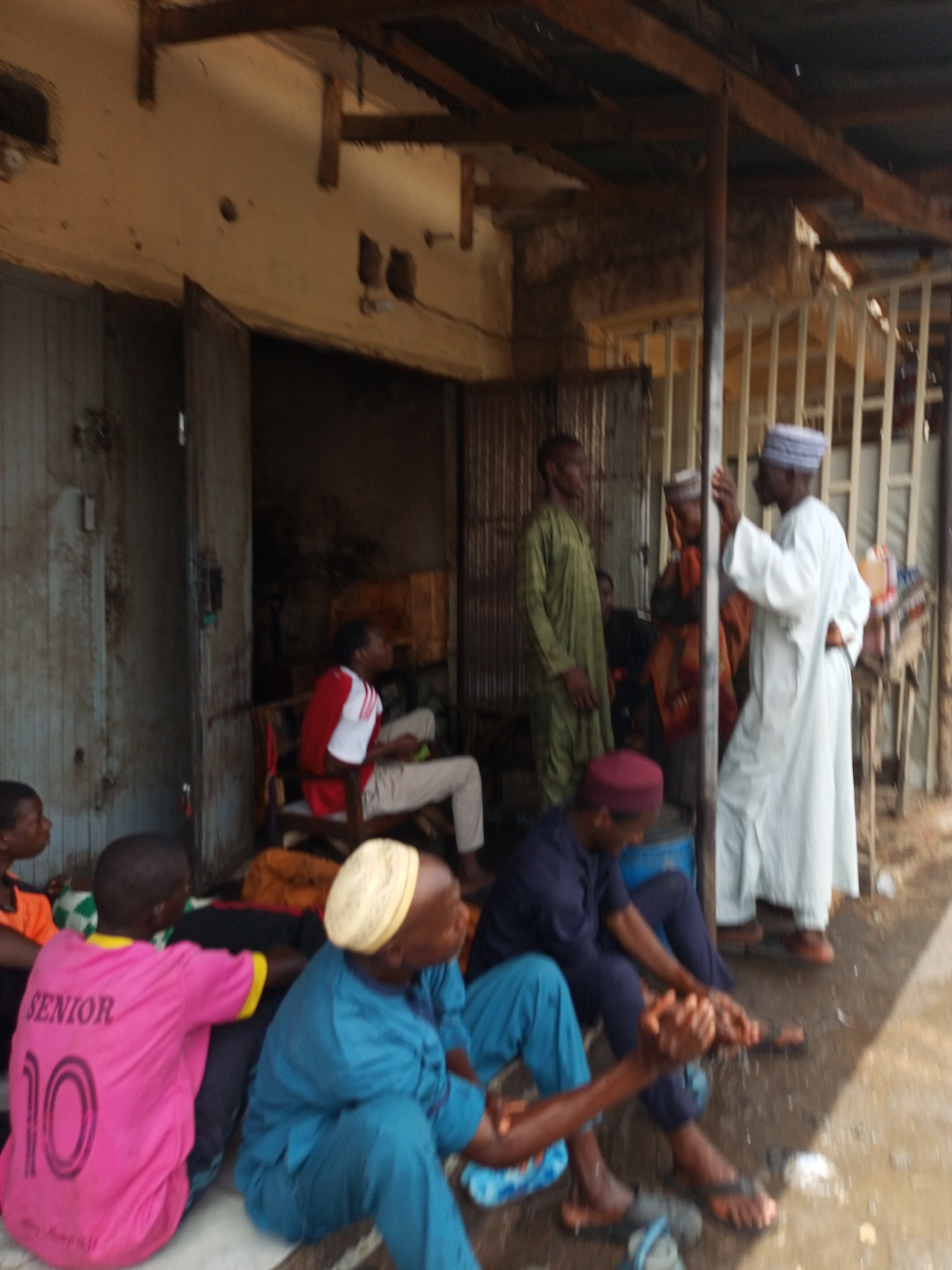 The image depicts a group of people gathered outside a weathered building with corrugated metal roofing in what appears to be an urban setting. Some individuals are seated on the ground, while others are standing and engaged in conversation. The men are wearing a mix of traditional and casual clothing. The environment appears somewhat dilapidated, with the building showing signs of age and wear. The location is indicated as Tempure, Nigeria.