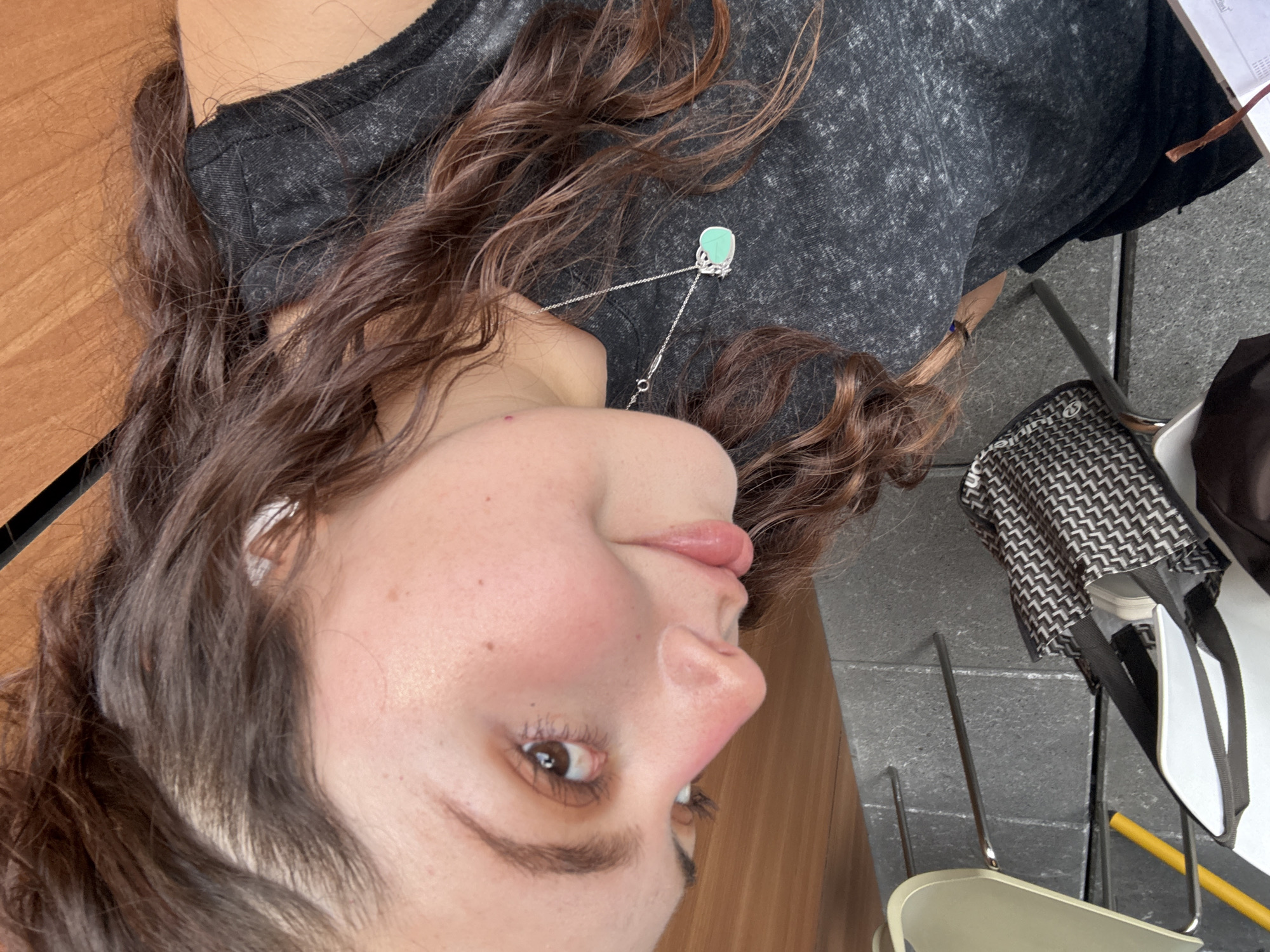 This media file captures a close-up, rotated view of a young woman situated in what appears to be a casual indoor or outdoor setting in Mexico City. The image is oriented sideways, with the woman's head facing towards the left side of the frame and her body extending upwards.
The primary subject is the woman, whose face, upper chest, and flowing hair are visible. She has fair skin with some freckles, light brown eyes, and full, slightly parted lips. Her expression is neutral and calm. Her long, wavy, dark brown hair cascades around her head and shoulders, blending with her dark gray, speckled t-shirt. A prominent silver necklace with a light green, possibly turquoise, square pendant hangs over her shirt, the chain visible extending towards her chest. A white earbud is nestled in her left ear.
The background shows elements of the setting. To the left and behind her head is a warm-toned wooden surface, likely a table. To the right, a darker, textured gray surface, possibly stone or concrete flooring, is visible. Several metallic chair legs extend from the bottom right, suggesting the presence of seating. A black and white patterned bag with a geometric design is seen resting on the gray surface near the chairs. Partial views of a notebook or papers with illegible text ("inal" visible) are at the top right, along with a yellow writing implement.
The image suggests a relaxed moment, possibly a casual photo or selfie. The lighting indicates daytime. No specific activity beyond posing is evident.