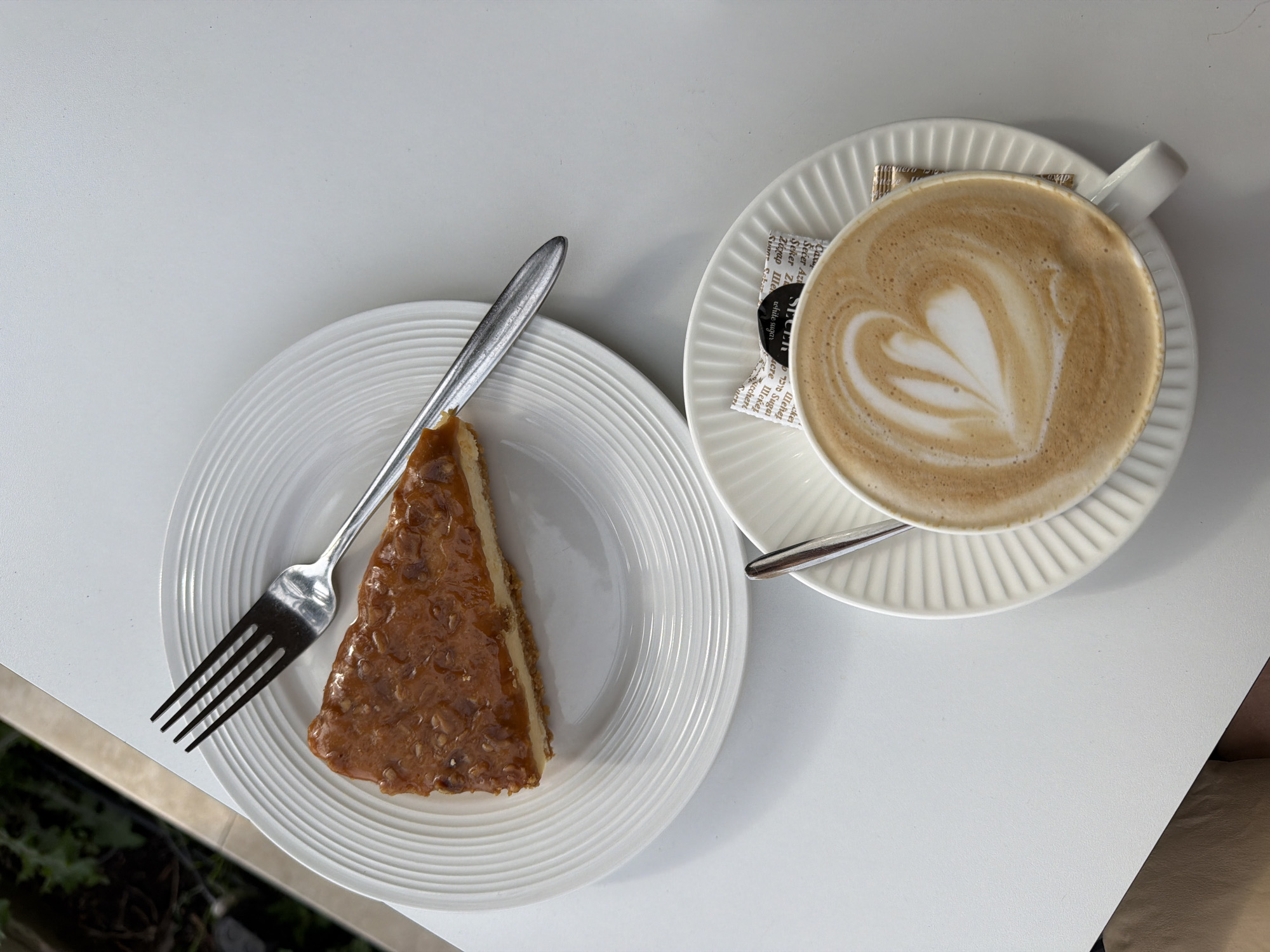 The image shows a top-down view of a white table setting with a piece of caramel cheesecake and a cup of latte art. The cheesecake, topped with a glossy caramel and nut mixture, rests on a white, ridged plate beside a silver fork. A cup of latte, showcasing a heart-shaped foam design, sits on a matching saucer accompanied by packets of sugar. A small silver spoon rests on the saucer. The table is white and appears to be outside, with hints of greenery in the lower left corner. No specific information within the image indicates that this is Bečići, Montenegro.