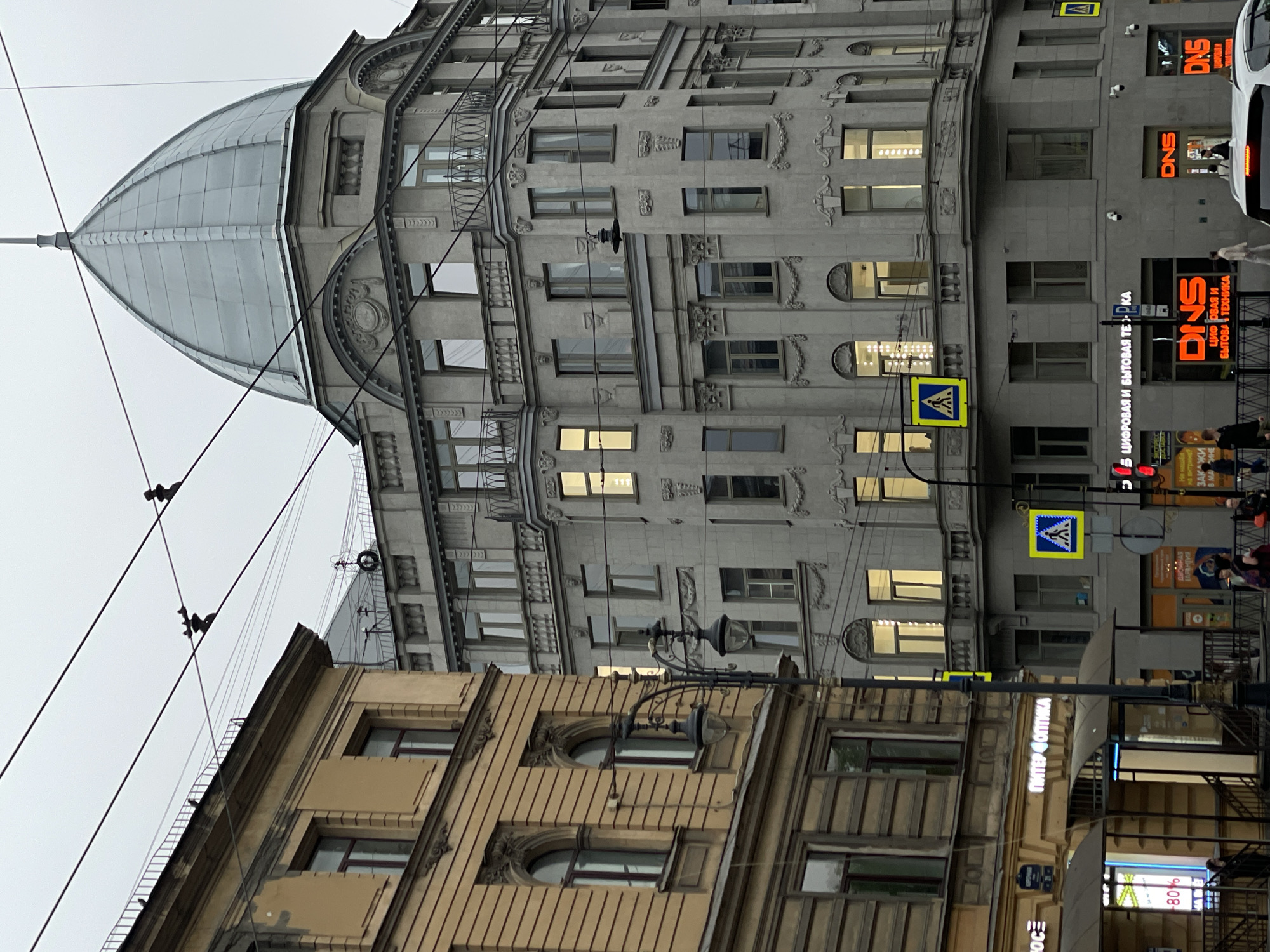 The image shows a street scene in Saint Petersburg, Russia. The buildings are made of gray and yellow stone. There is a large dome on top of one of the buildings. There are also some utility poles and wires in the foreground, with bird diverters strung along the wires. There are pedestrian crossing signs near streetlights. Storefronts for businesses such as "DNS" (a digital and household appliance company), and "Питер Оптика" ("Peter Optica") are visible. There are people walking on the sidewalk. Part of a white vehicle is visible on the right side of the image.