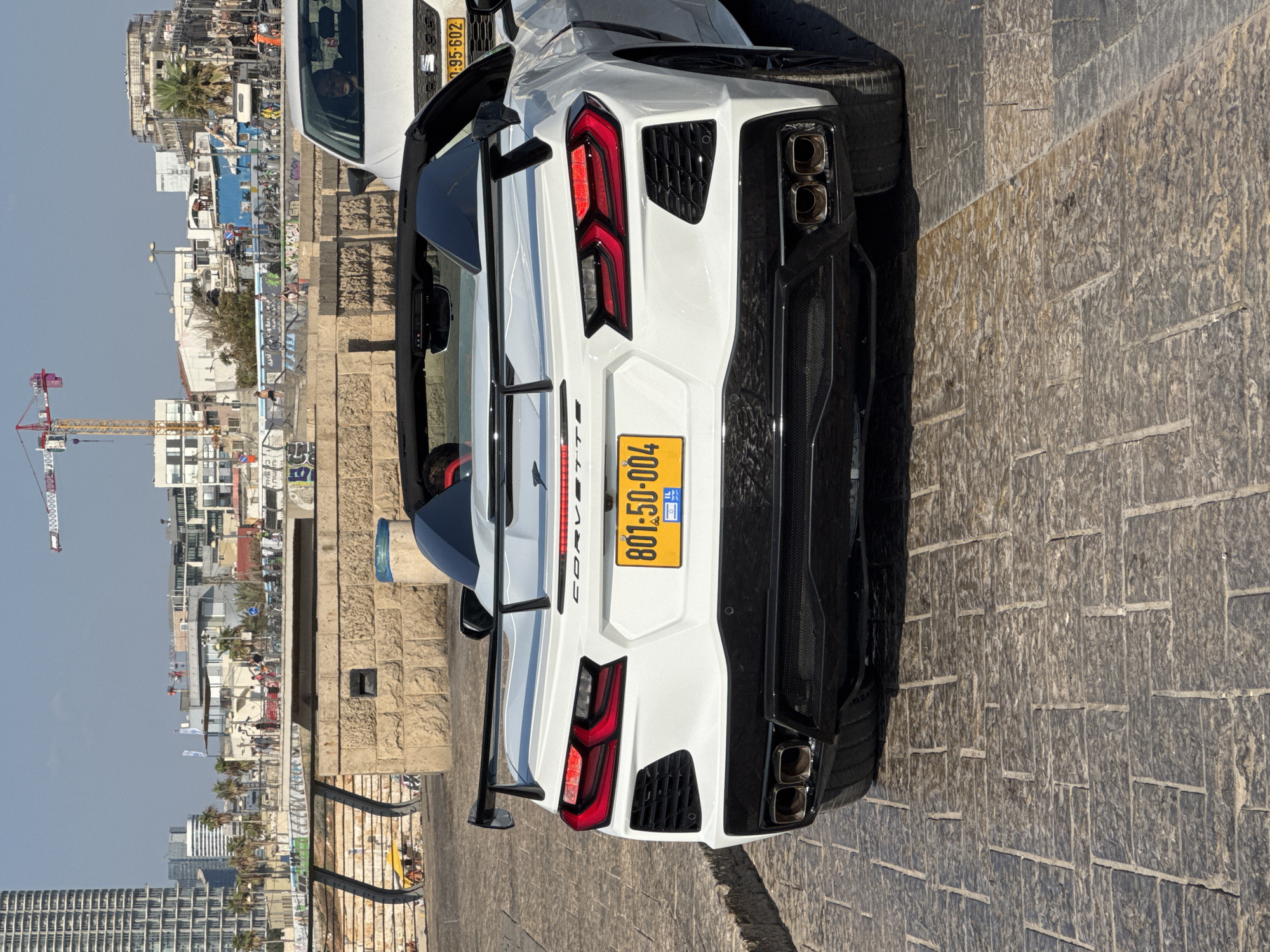 The image captures the rear view of a white Chevrolet Corvette C8 convertible parked on a cobblestone street. The car features a prominent rear spoiler, distinctive taillights, and quad exhaust tips. The license plate, which reads "801-50-004 IL," is yellow with black lettering, indicating it is an Israeli plate. To the right, a white SUV is partially visible, with its license plate showing "95-602." In the background, a cityscape unfolds with modern buildings, palm trees, a construction crane, and a body of water, suggesting a coastal urban setting. The scene appears to be along the Tel-Aviv promenade or a nearby street, given the architectural style and urban elements, characteristic of the city. People are visible in the mid-ground, strolling along a walkway or pier, and some are seated on a stone structure. The overall atmosphere is bright and sunny, with clear blue skies.