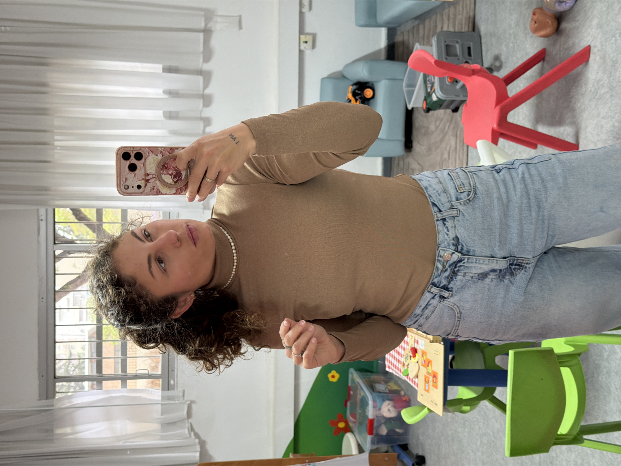 A woman is taking a selfie in a room containing children's furniture and toys. The room is located in Kiryat Bialik, Israel. The woman is wearing a brown turtleneck sweater, jeans, and a pearl necklace. She is holding a cell phone with a floral case in her right hand. The background includes a window with sheer curtains, a blue child-sized chair, a red toy chair, a green toy table, and various toys.