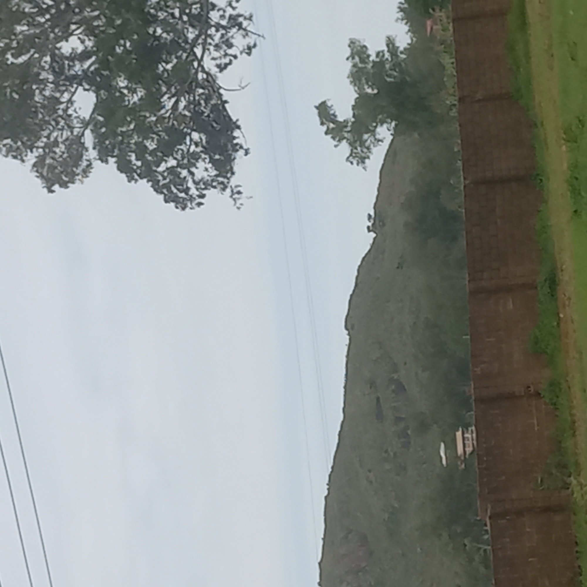 The image shows a landscape with a brick wall in the foreground and a hilly terrain in the background. A tree with dark foliage is visible on the right side of the image, partially obscuring the sky. In the middle ground, a building with multiple levels can be seen atop the hill. The sky is overcast and light gray. The location is identified as Kuru Dungus, Nigeria.