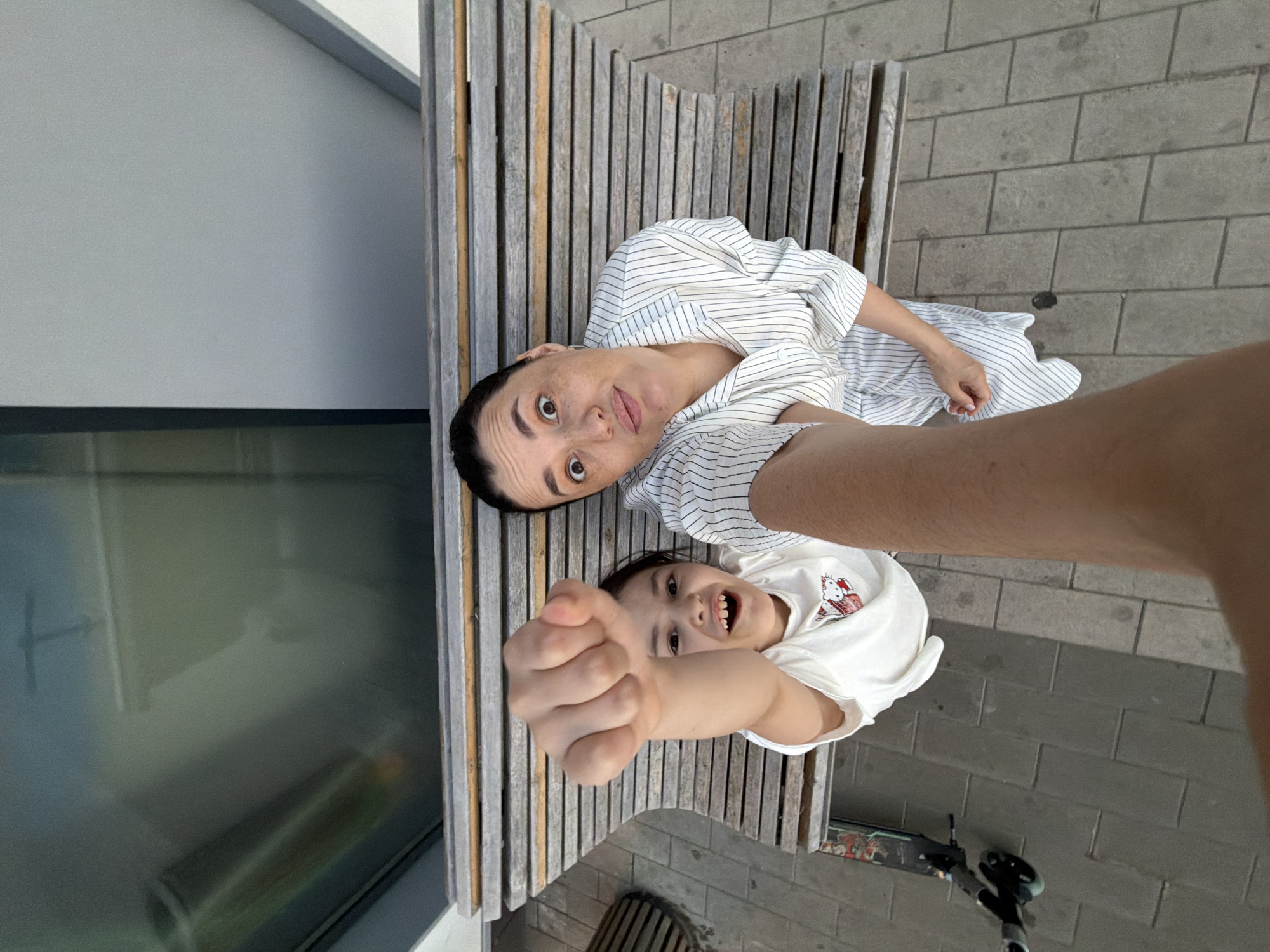 A high-angle, close-up, selfie-style shot captures a woman and a child sitting on a slatted wooden bench, likely outdoors. The woman, positioned on the right, wears a white shirt with thin black pinstripes and has an expressive face with wide eyes and freckles. The child, on the left, looks directly at the camera with a wide smile and a fist raised. A hand and arm, presumably belonging to the photographer, are visible in the foreground, holding the camera. In the background, a window reflects a distorted image of what appears to be an indoor structure. The ground is paved with gray rectangular tiles, and to the left of the bench, a black electric scooter is parked. The overall lighting suggests daylight. The specific location of Moscow, Russia, cannot be determined from the visual information provided in the image.