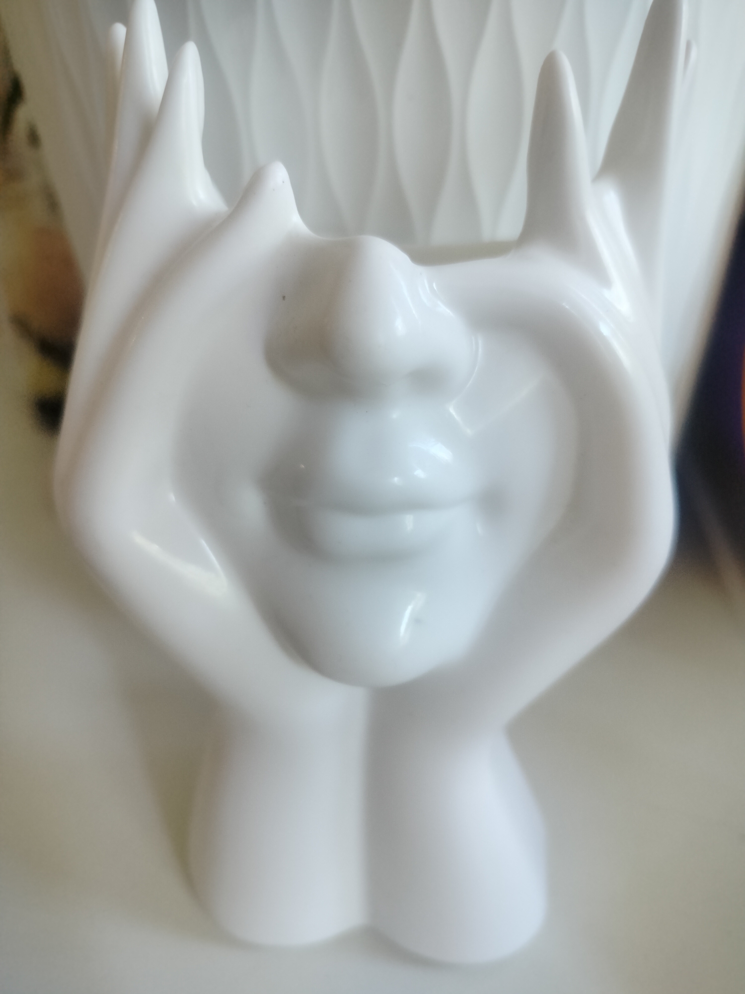 The image is a close-up, eye-level shot of a white ceramic vase shaped like a stylized face. The face has hands covering its eyes, creating a playful and somewhat secretive expression. The lips are slightly parted, and the nose is subtly defined. The vase itself has a glossy finish, reflecting the light and highlighting its smooth curves. The background is out of focus, showing hints of a light-colored, textured surface that might be a wall or another decorative item. There are also some blurred colors in the far background, possibly from other objects or decorations. The lighting appears to be artificial and soft, creating gentle shadows and highlights. The overall impression is one of artistic craftsmanship and a whimsical design. There is no visible text in the image.