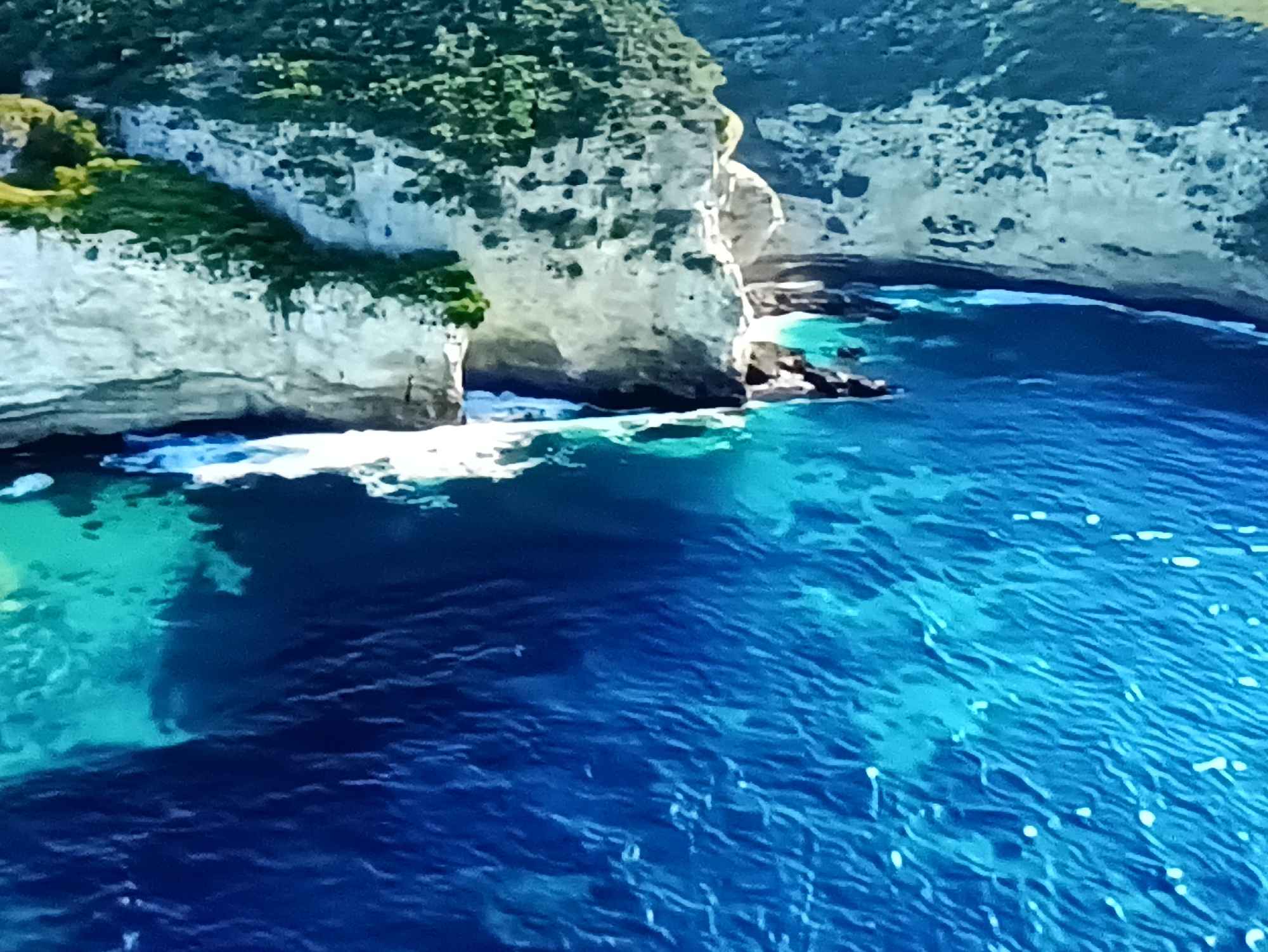 This is a high-angle, aerial view of a dramatic coastal landscape featuring towering, rugged cliffs meeting the ocean. The cliffs are primarily light-colored, with patches of green vegetation clinging to their surfaces, suggesting a vibrant ecosystem despite the harsh terrain. The ocean itself is a striking feature, transitioning from shallow, turquoise waters near the shore to a deep, rich blue further out. White waves crash against the base of the cliffs and break into foam, highlighting the powerful interaction between the sea and land.
The scene depicts a natural, undeveloped location, emphasizing the raw beauty of the coastline. The water is clear enough in some areas to reveal the rocky seabed below, adding to the visual depth. Sunlight reflects off the water's surface, creating shimmering patterns and bright highlights. The overall impression is one of a remote and pristine natural environment, likely experienced on a clear, sunny day given the bright illumination and vibrant colors. There is no visible text in the image. The location context provided, Kamianets-Podilskyi, Ukraine, is unusual for this type of coastal scenery, as Kamianets-Podilskyi is an inland city known for its castle and historical architecture, not its coastlines. This suggests the image may be misattributed or represent a different location than stated.