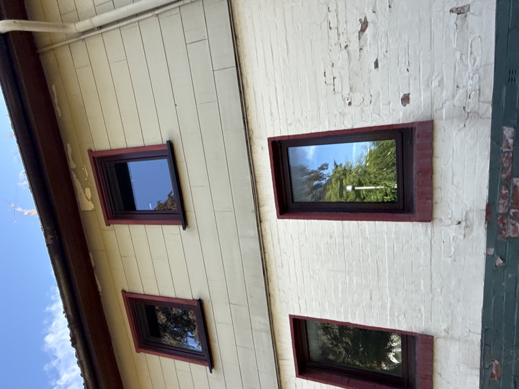 This is an outdoor, eye-level, medium shot of the side of a building in Walkenried, Germany. The building has two distinct levels. The upper level is clad in light beige horizontal siding. It features two dark-framed windows, each with a dark red trim. The lower level is constructed of white-painted brick, and also has dark-framed windows with dark red trim. A dark green band runs along the bottom of the brick wall. The sky above is blue with white clouds. Reflections in the windows show trees and the sky. The overall impression is of a residential building.