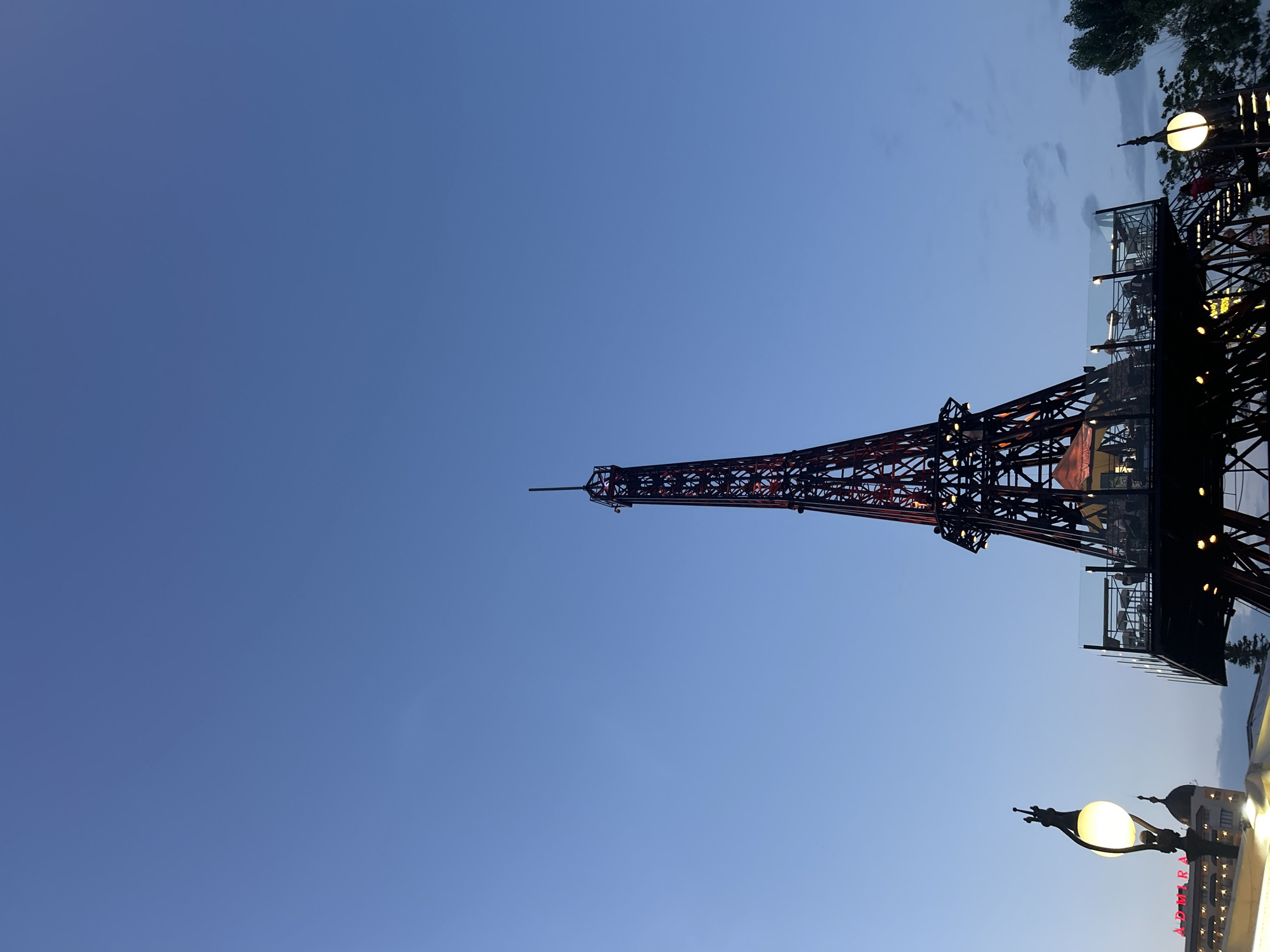The image presents a vertical, eye-level shot of a darkened, miniature Eiffel Tower replica standing against a twilight sky, identified as being in Varna, Bulgaria. The sky transitions from a lighter blue at the top to a deeper, purplish-blue near the horizon, with faint, wispy clouds visible in the lower right quadrant. The tower structure is made of dark metal latticework, illuminated by sparse, reddish-orange lights embedded within its framework, suggesting it is operational as a commercial establishment. A multi-level platform, serving as an outdoor restaurant or bar, is integrated into the lower section of the tower, featuring tables, chairs, and glass railings. Several indistinct human figures are visible on these platforms, indicating patronage. To the left of the tower, a tall, ornate street lamp with a glowing spherical white light is prominent. Behind the street lamp, a building with a rounded, dome-like roof and a partial red neon sign reading "AD MIRA" is visible. On the far right, a section of a tree with green foliage extends into the frame. The scene is illuminated by a combination of fading natural light and artificial lighting from the tower, the street lamp, and the building sign.