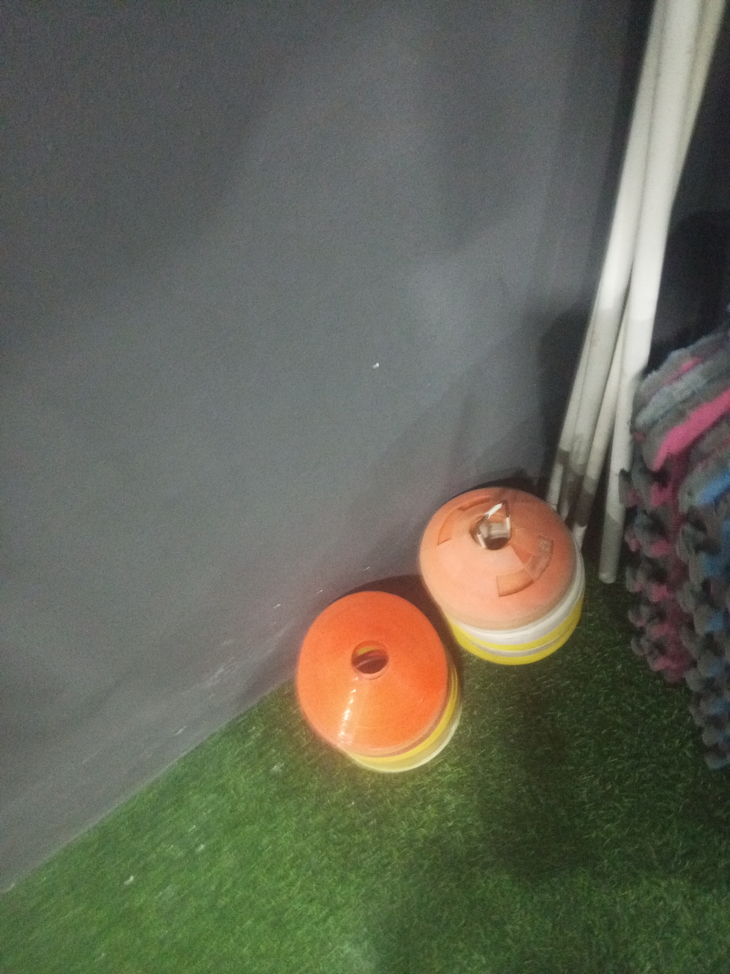 The image depicts a corner of an indoor space, likely a gymnasium or sports facility, with a dark gray wall to the left and a green artificial turf floor. In the foreground, two stacks of orange and yellow training cones are visible. Behind the cones, leaning against the wall, are several long, thin, white cylindrical objects, possibly poles or floor markers. To the right of these, a stack of folded mats or pads, in shades of pink, blue, and gray, is propped up. The lighting is dim, casting shadows from the objects onto the wall and floor. There are no people or animals present in the scene. The overall impression is one of stored sports equipment. The location of Juan Griego, Venezuela, is not indicated by any visual elements within the image itself.