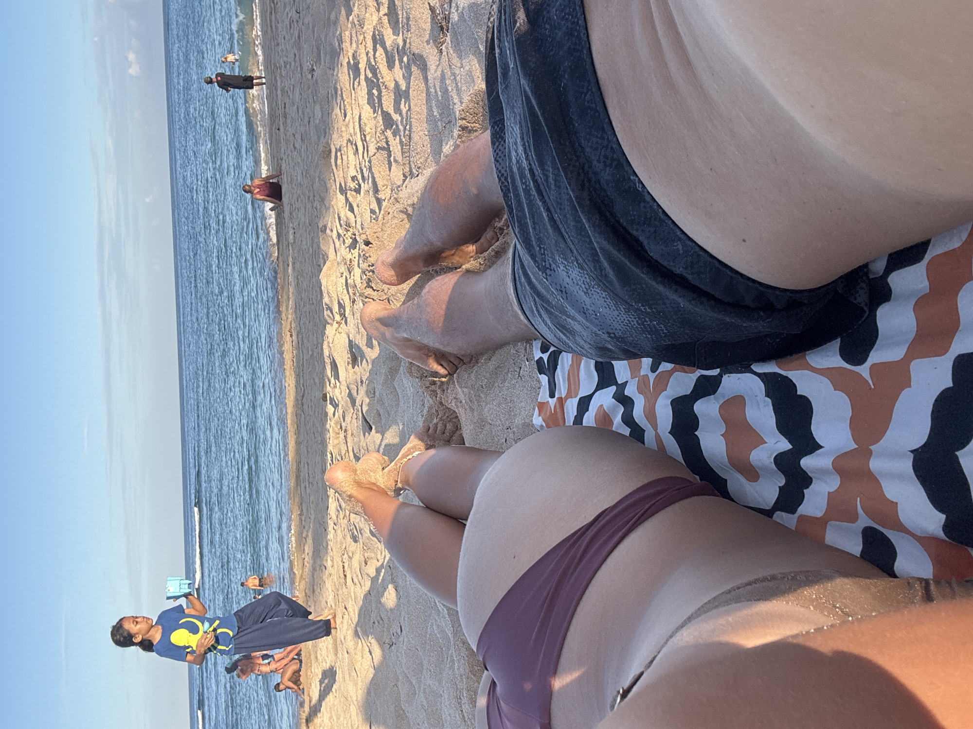 The image shows a sandy beach scene, potentially near Kuta Selatan, Indonesia, given the beach and ocean setting. Two people are lying on a patterned beach towel. The person in the foreground is wearing a dark purple bikini bottom. The other person is wearing dark shorts. The edge of the ocean and several people in the distance are visible in the background. A child is standing on the beach playing with a water pistol.