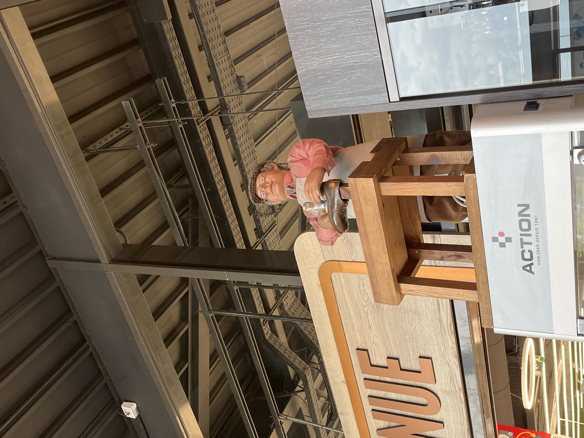 This is a medium shot, eye-level, outdoor, full-body photograph of a mannequin in a store in Le Port-Marly, France. The mannequin is depicted as a shoemaker, wearing a red and white checkered shirt, an apron, corduroy pants, brown shoes, and a flat cap. It is positioned on a wooden platform, holding a shoe in its hands as if repairing it.
Behind the mannequin, a wooden sign with orange trim displays the word "NUE" in large, light brown letters. The sign appears to be part of the store's facade.
In the foreground, a white structure with the "ACTION HORLOGER DEPUIS 1961" logo is visible. To the right of the mannequin, a dark grey, metallic structure with a glass panel reflects the sky.
The background consists of a corrugated metal ceiling structure, crisscrossed with metal beams and conduits, suggesting an indoor or partially covered area of the store. The lighting is bright, indicating daytime. The overall scene conveys a commercial setting, likely a retail store specializing in shoes or watches, given the "ACTION HORLOGER" branding.