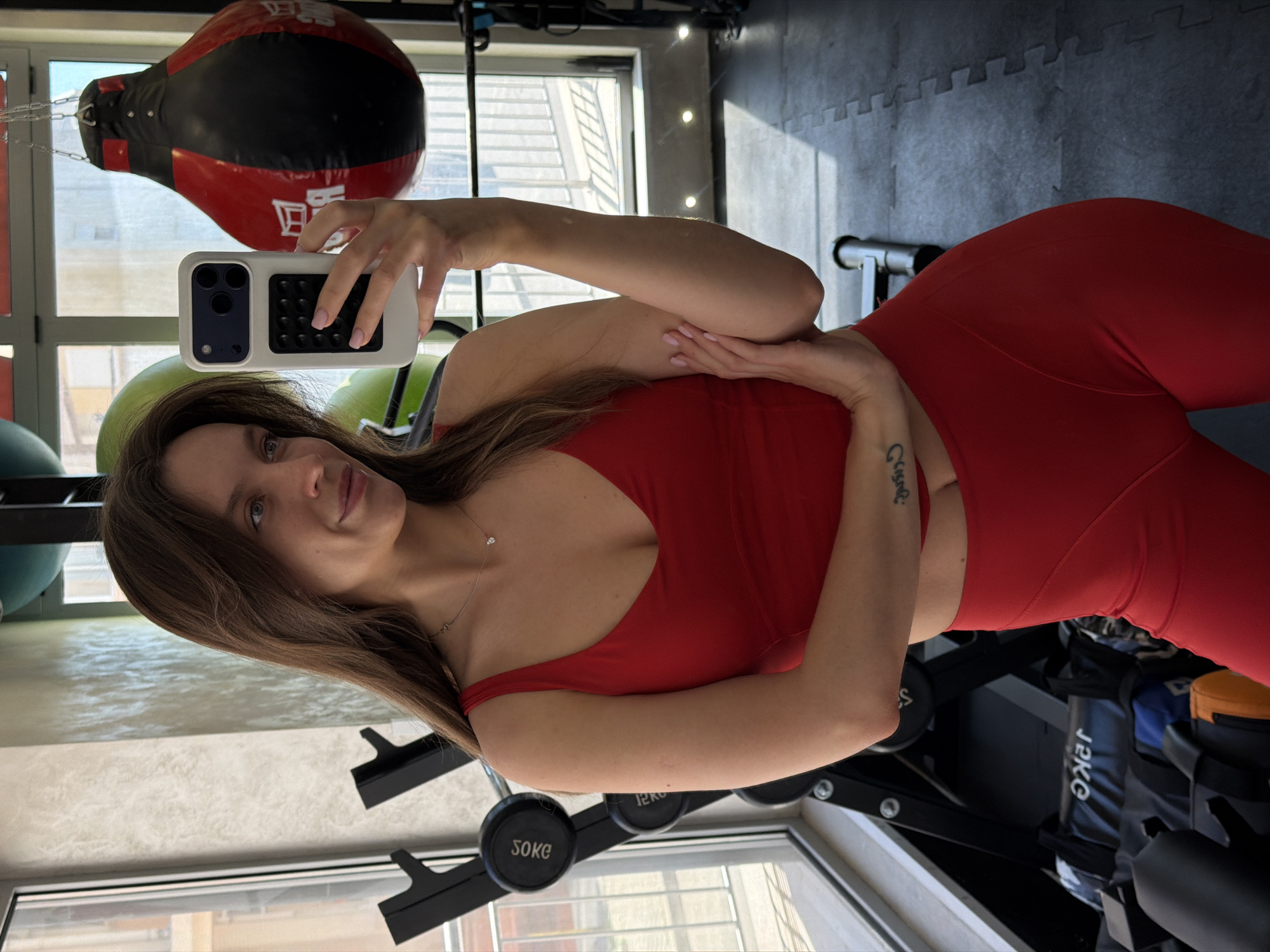 A young woman is featured in a selfie taken indoors, likely within a gym or fitness studio. The image is oriented 90 degrees clockwise, making her appear to be standing and looking towards the camera. She has long brown hair, fair skin, and light-colored eyes, displaying a subtle, pleasant smile. She is dressed in a form-fitting red athletic top, possibly a sports bra, and matching high-waisted red leggings. A delicate silver necklace with a small pendant is visible around her neck, and a tattoo, appearing to read "Crise" or similar, is present on her right forearm.
She holds a white smartphone with a distinctive black camera module and a black textured grip on the back. The background reveals typical gym equipment: a red and black punching bag hangs in the upper left, while green exercise balls are partially visible. Various gym machines and weights, including a barbell rack with "50KG" and "SOKE" labeled on weight plates, are scattered around. The floor is covered with black interlocking mats. Natural light streams through large windows, indicating a daytime setting. A "JKKC" branded item is also visible near the bottom right. The scene depicts a typical gym environment, consistent with a fitness center in a location like Budva, Montenegro.