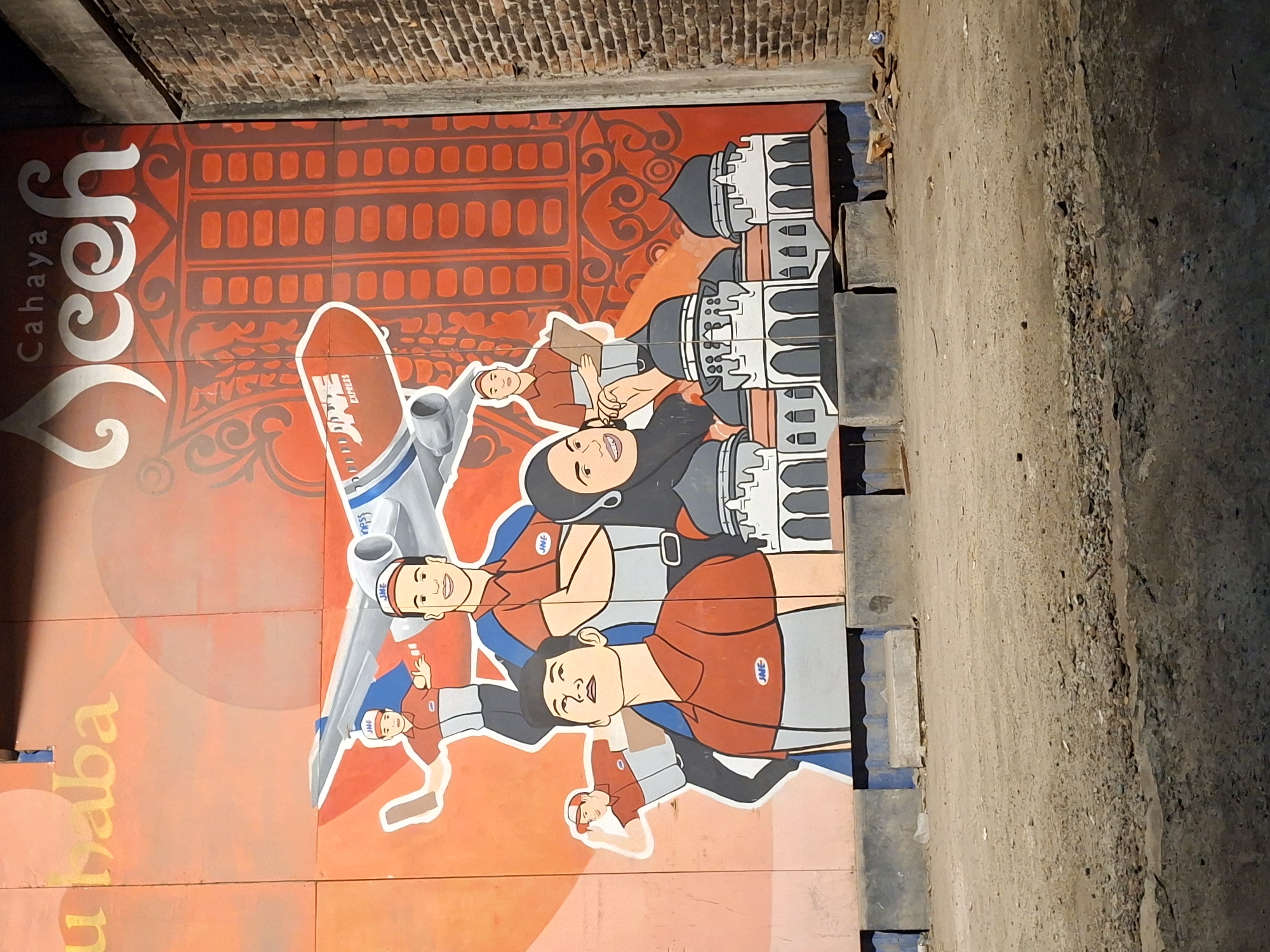 The image depicts a vibrant mural in Lhokseumawe, Indonesia. The mural is painted on a section of wall that appears to be part of a bridge or underpass. The artwork features cartoon-like figures, including men in JNE Express uniforms and a woman in a hijab. There is also an airplane and depictions of architectural structures that are likely relevant to the local culture or history. The background is predominantly orange and red with decorative patterns. The wall is flanked by a textured, concrete-like surface on one side and a brick wall on top.
