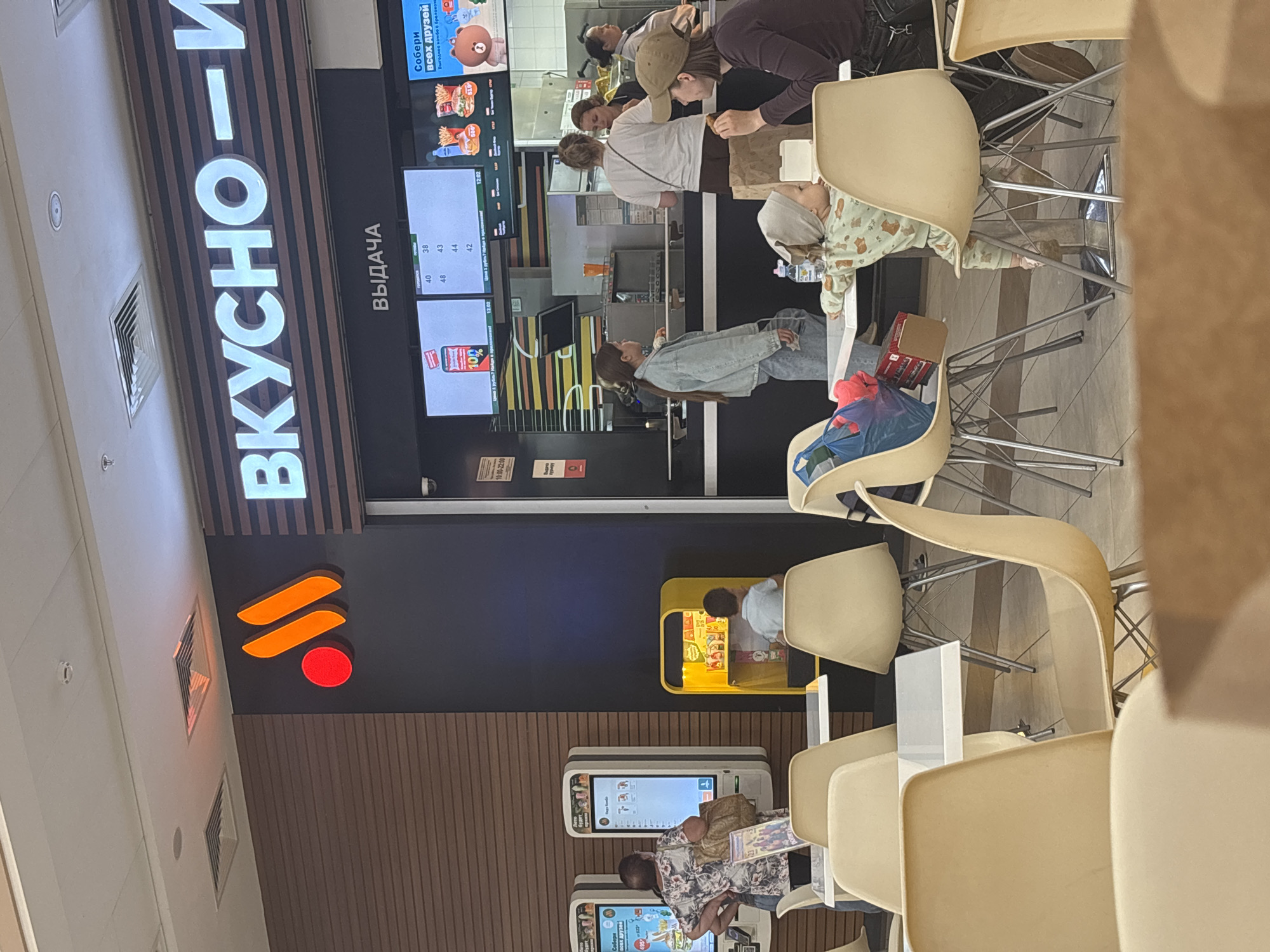 In an indoor fast-food establishment, possibly a McDonald's given the branding and menu displays, several individuals are visible. A baby in a light green onesie sleeps peacefully in a high chair, its head resting on a gray pillow. Nearby, a woman in a denim jacket is seated at a table, looking towards a digital menu. Another woman, wearing a patterned shirt, is also seated, possibly waiting for food. In the background, a few other people are engaged in ordering or dining.
The scene is illuminated by artificial lighting, suggesting it could be any time of day. The setting appears to be a modern, clean fast-food restaurant with light-colored seating and tiled floors. Large digital screens display menu items and promotions. One screen shows a "10% Off" offer. Another screen displays prices like "40", "38", "48", "43", "44", and "42", along with a time stamp of "12:02". A large illuminated sign in Russian reads "ВКУСНО - И", which translates to "TASTY - AND". Another sign says "ВЫДАЧА", meaning "ISSUE" or "DISPENSING". A prominent logo features stylized orange swooshes and a red circle, reminiscent of the McDonald's branding. The overall atmosphere is casual and functional, typical of a fast-food dining area. There is also a child-friendly play area visible with yellow panels. The text "Собери всех друзей" (Collect all friends) and "Выгодное комбо с брелоком" (Profitable combo with a keychain) indicates a promotional campaign. The time "10:00-22:00" suggests operating hours.